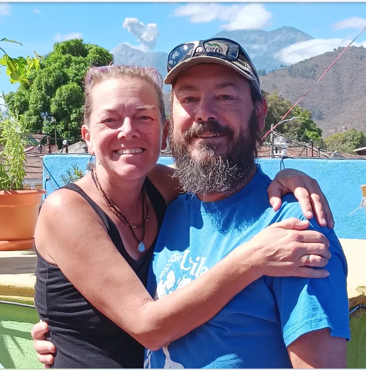 Two people smiling outdoors, one with arm around the other, mountains and blue sky in background.