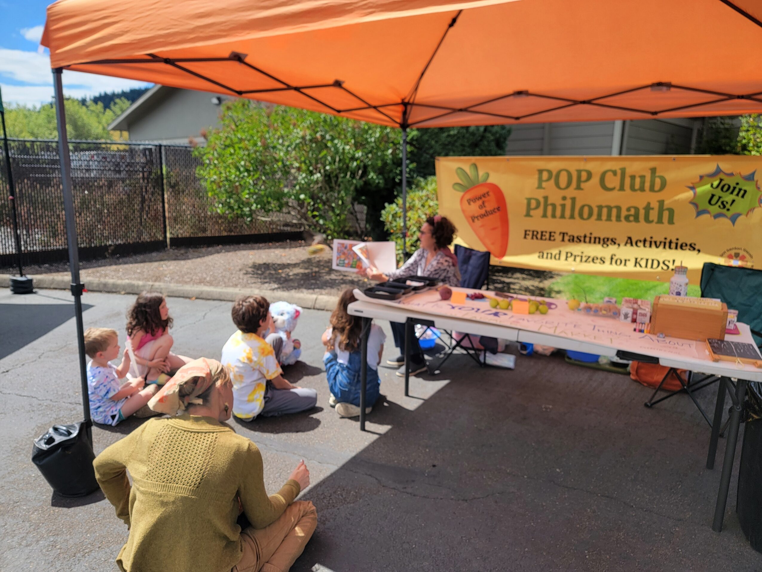 Children sitting on the ground in front of a table under an orange canopy at a community event.