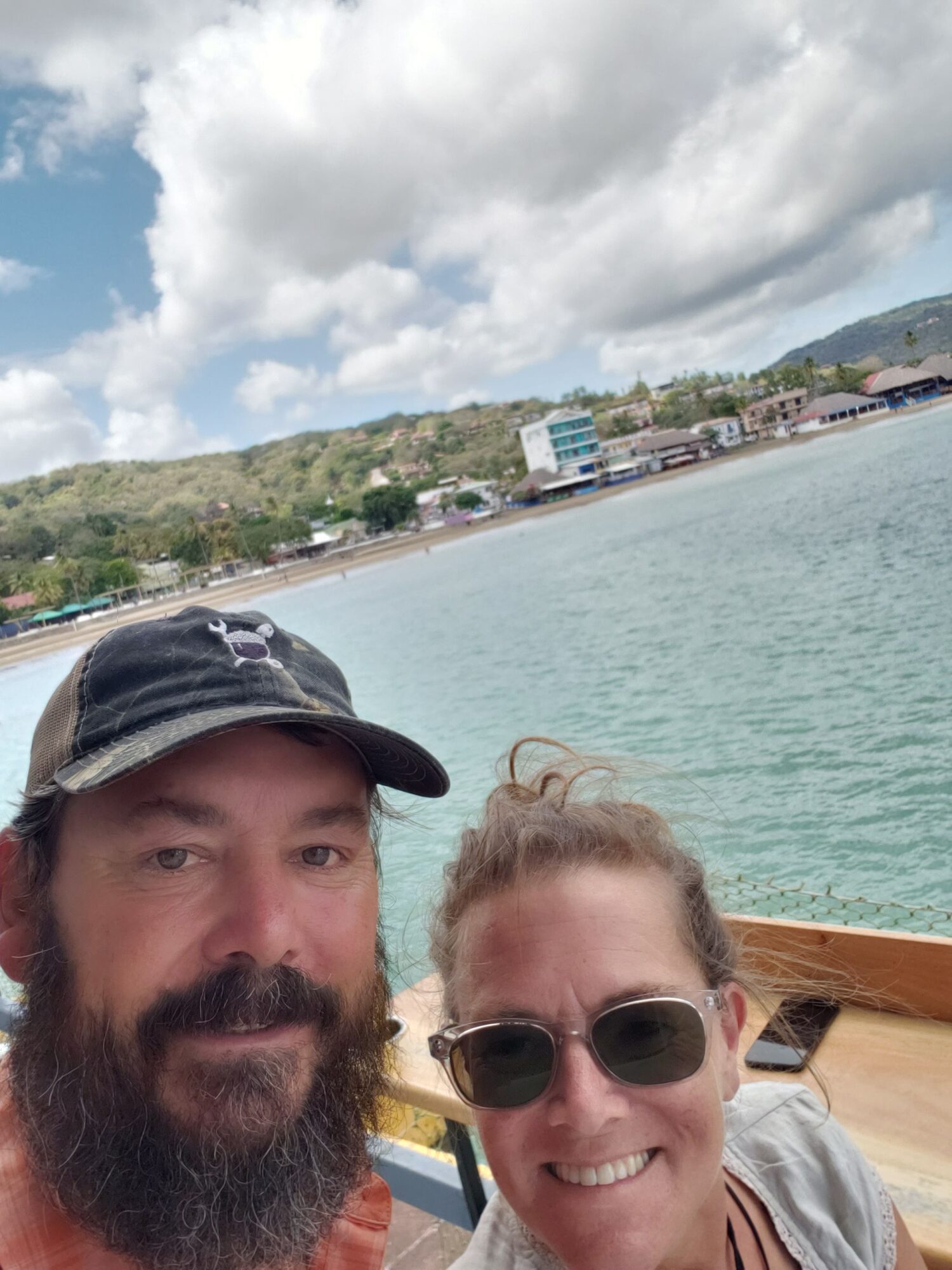 Two people smiling near water with a cloudy sky and shoreline in the background.