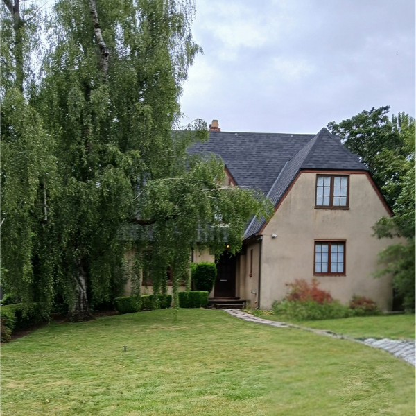 House with a sloped roof, surrounded by trees and a lawn, viewed from the front yard.
