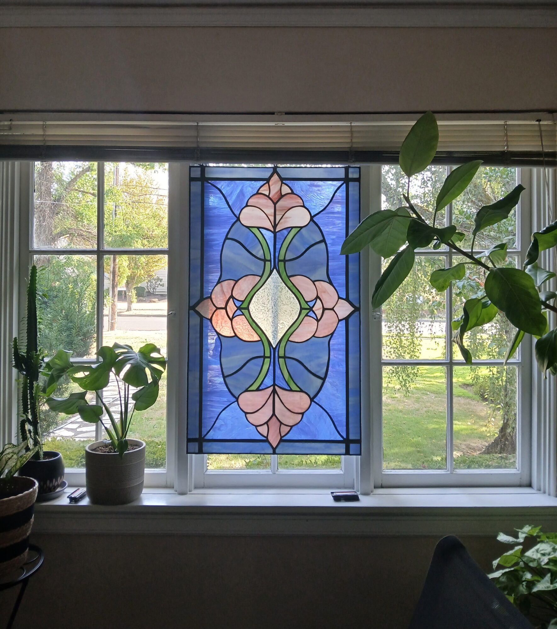Decorative stained glass window with floral design, flanked by potted plants on a windowsill, with a view of trees outside.