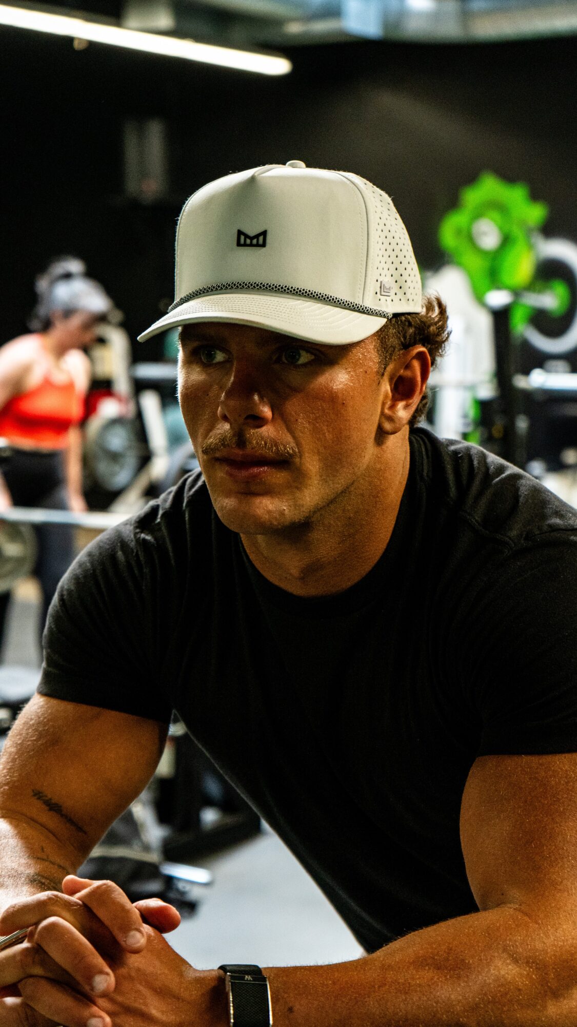 Man wearing a white cap and black shirt, sitting indoors with a blurred background, including a person in red and green gear.