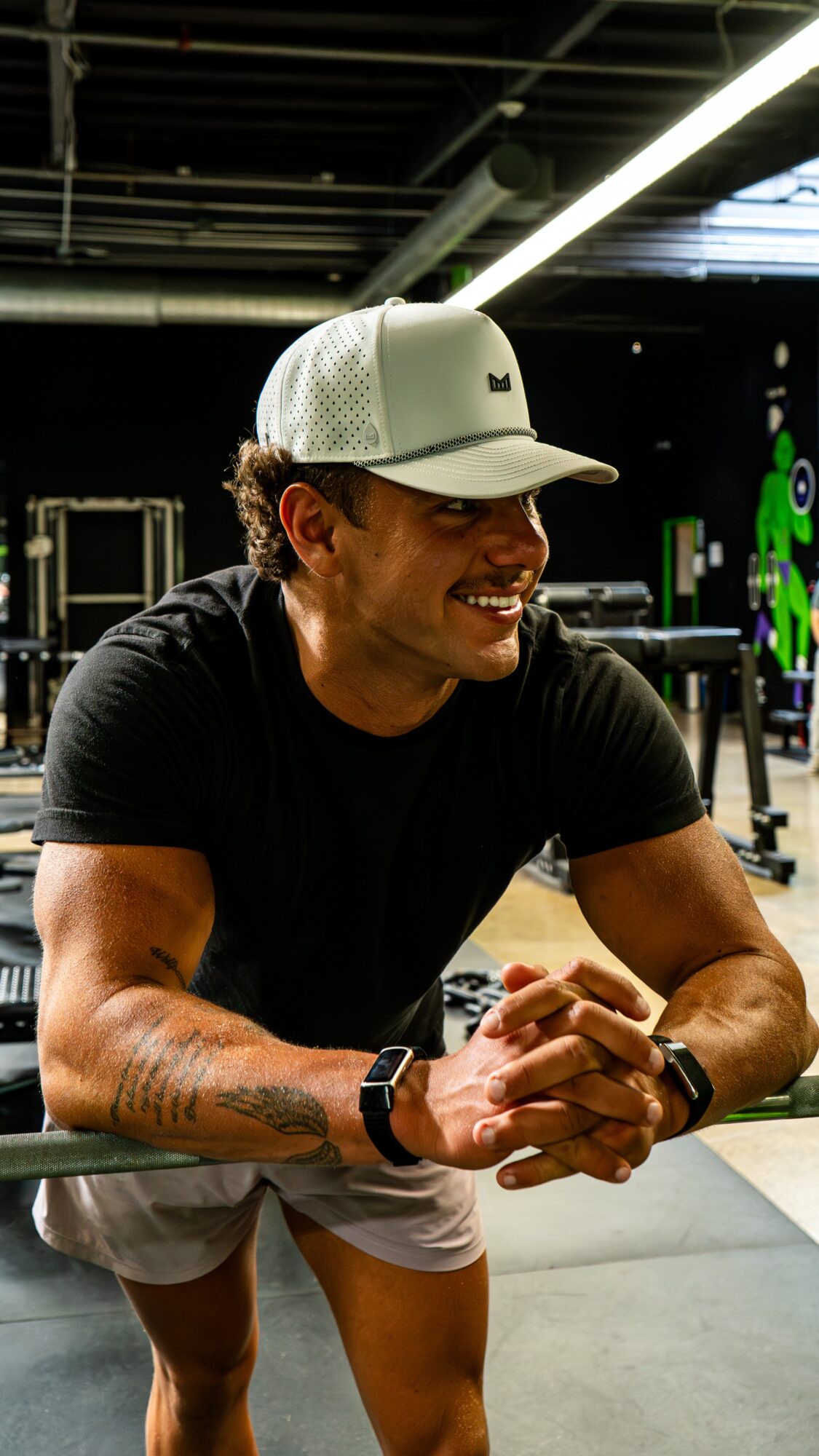 Young man smiling, wearing a white cap and black t-shirt, sitting in a gym with equipment in background.