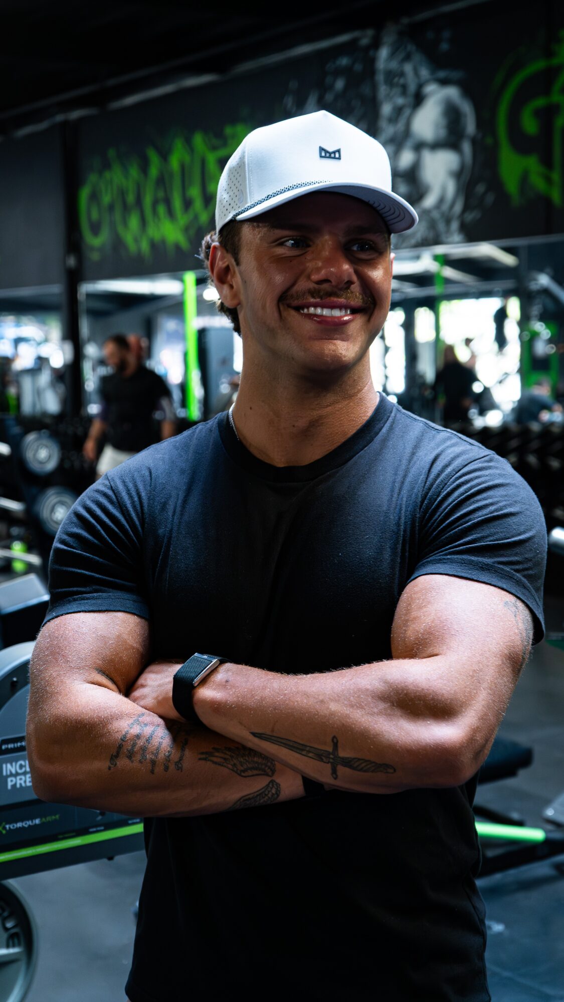 Smiling man with crossed arms wearing a white cap and black shirt in a gym, with green signage in background.