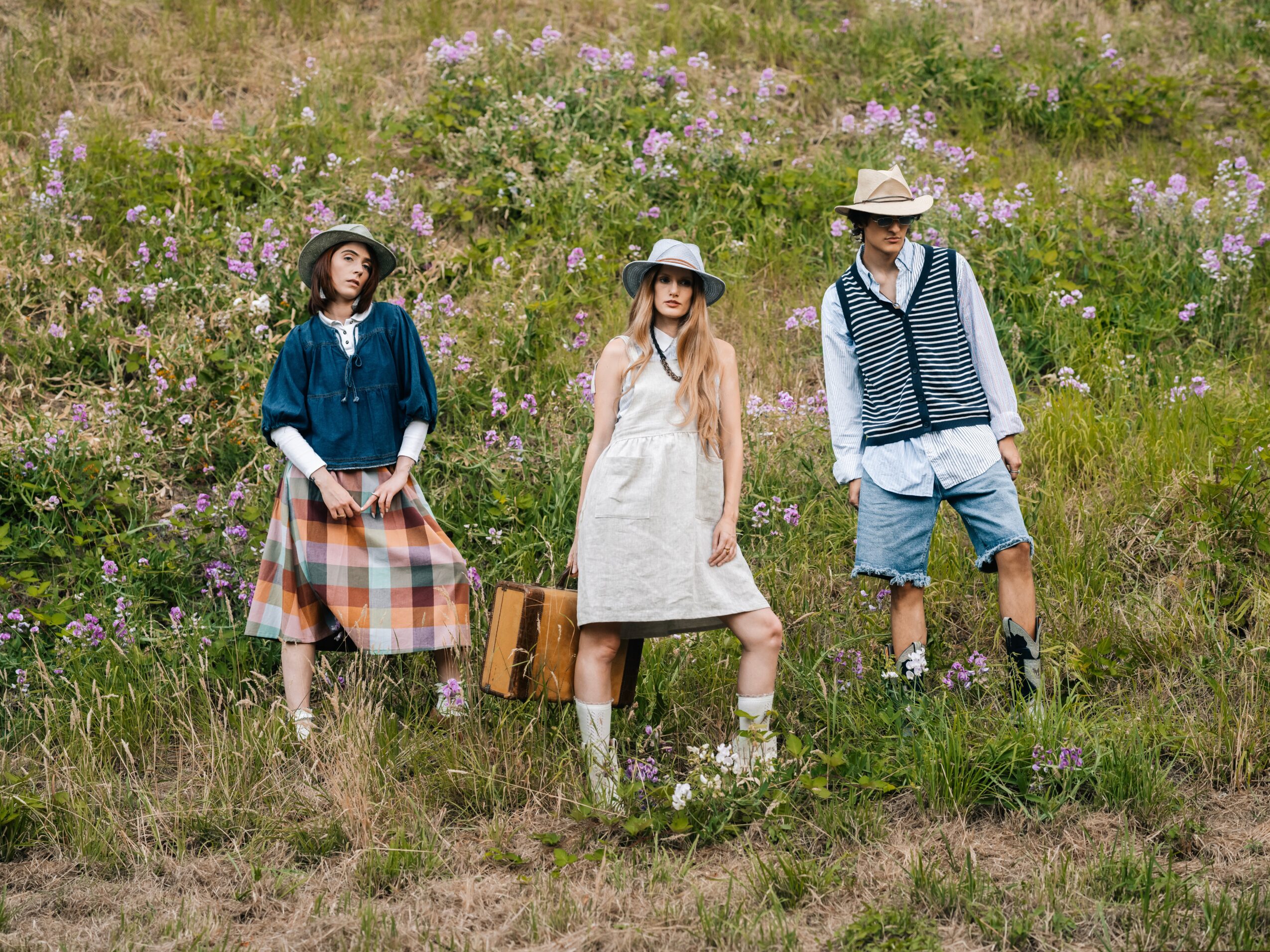 Three people stand in a grassy field with purple flowers, wearing summer clothing and hats, facing forward.
