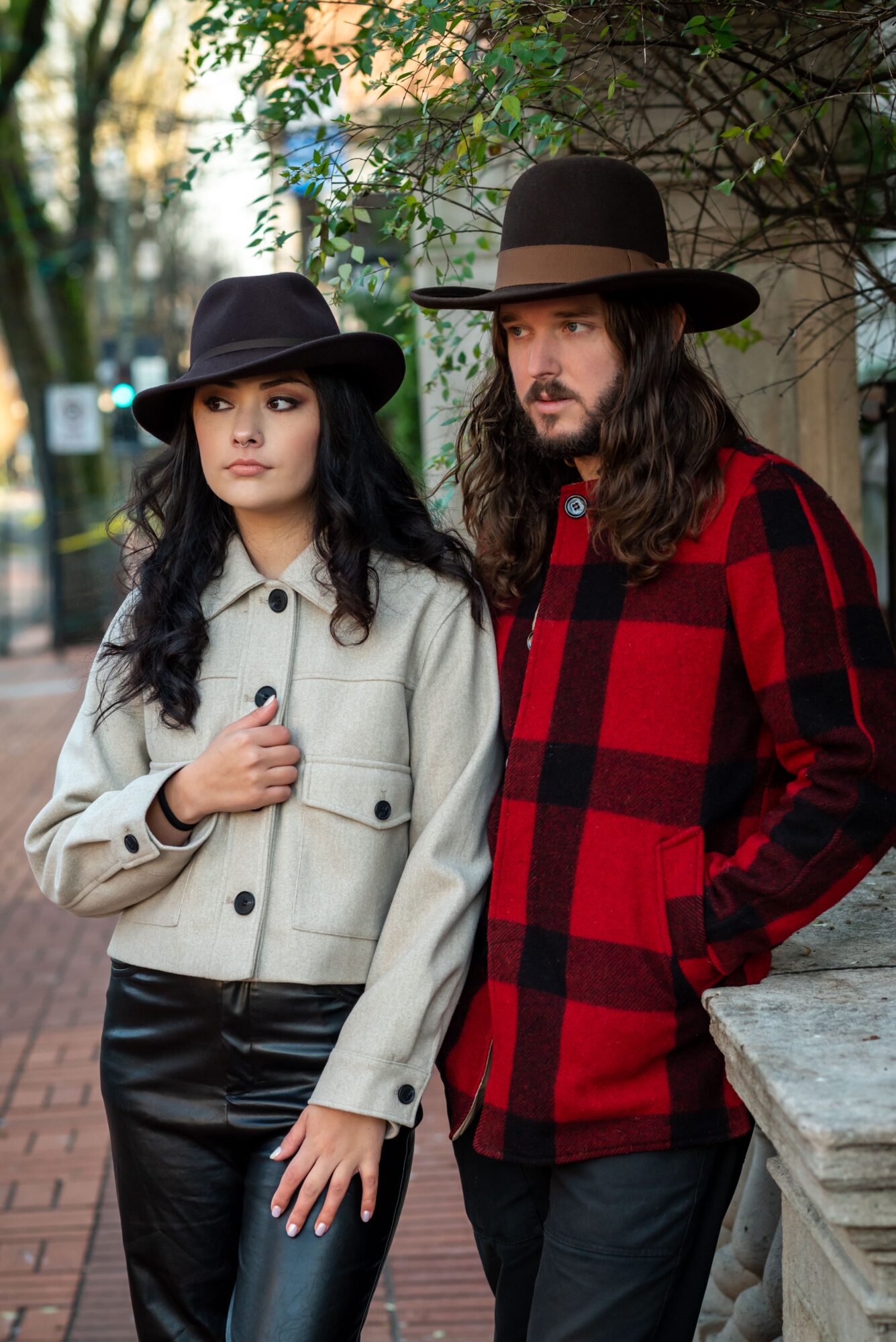 A woman and a man stand outdoors on a brick path, with trees and a stone wall behind them.