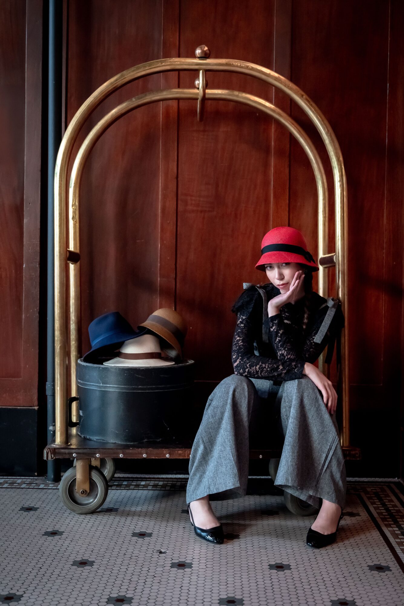 Woman sitting on a luggage cart with hats, wearing a red hat and gray wide-leg pants, against a wooden wall.