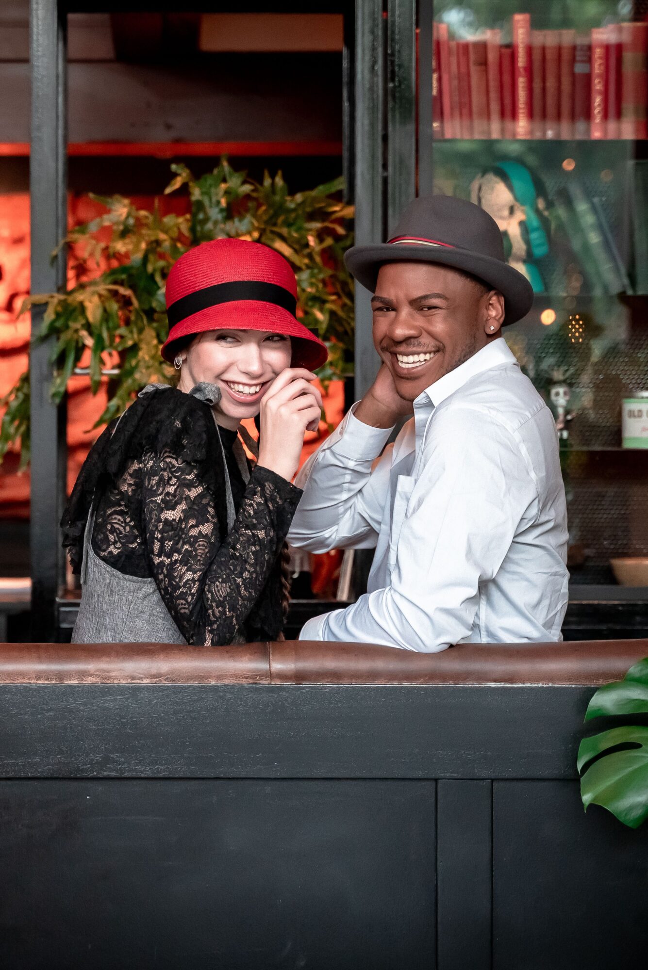 Two people smiling and laughing, one wearing a red hat and the other a gray hat, sitting at a table in a cozy setting.