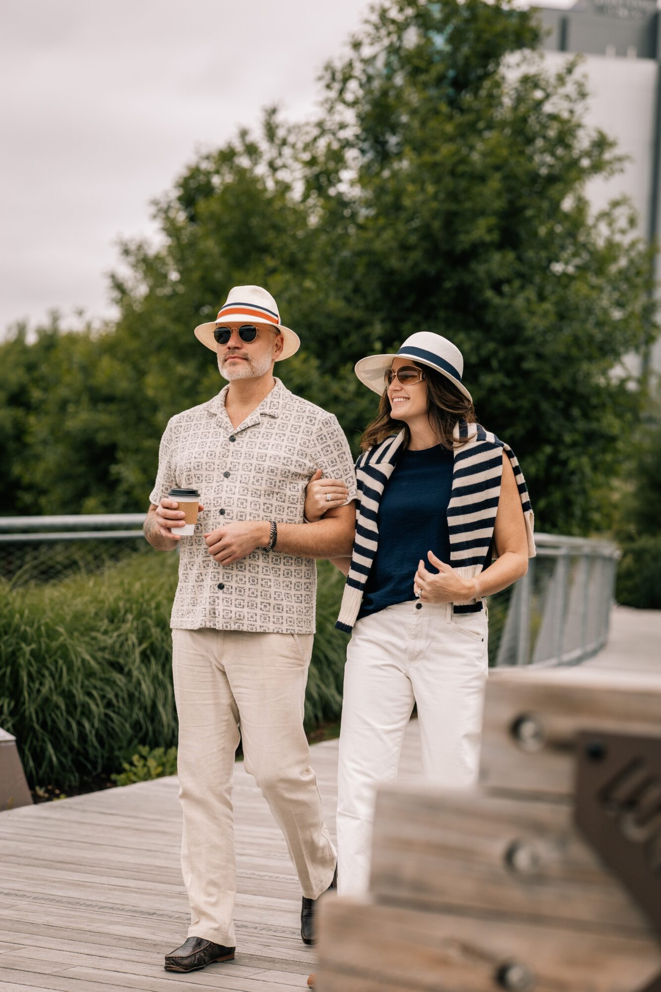 A man and woman walk outdoors on a wooden path, holding drinks, with trees and a railing in the background.