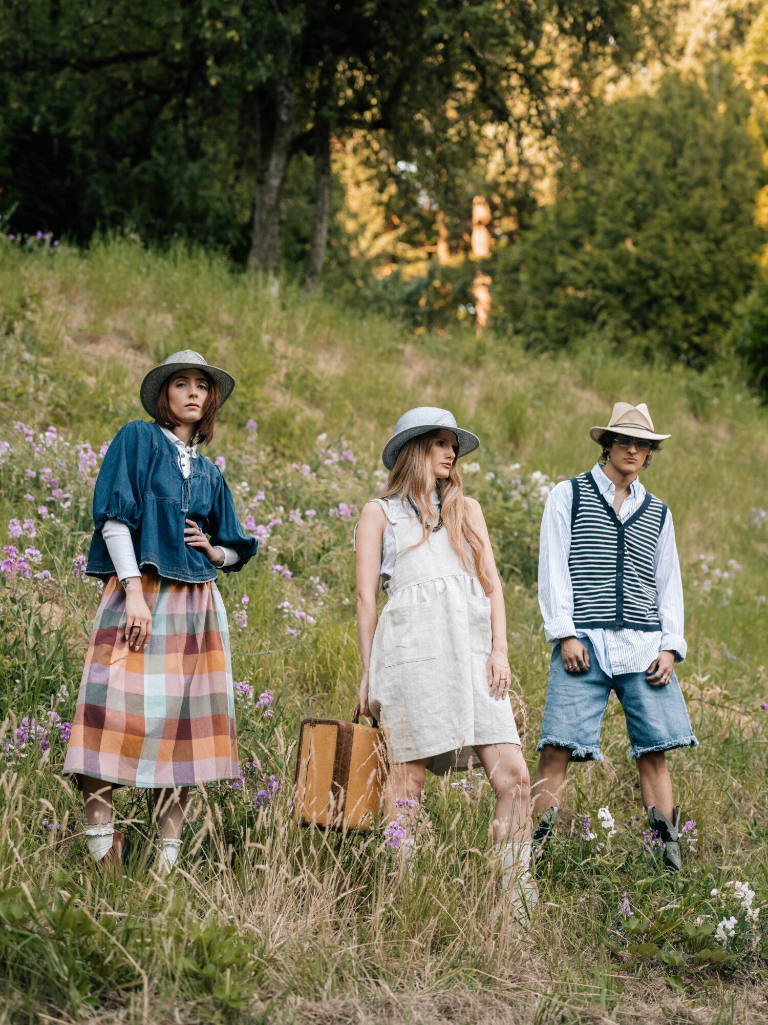 Three children standing in a grassy field with trees and flowers, wearing summer clothing and hats, during sunset.