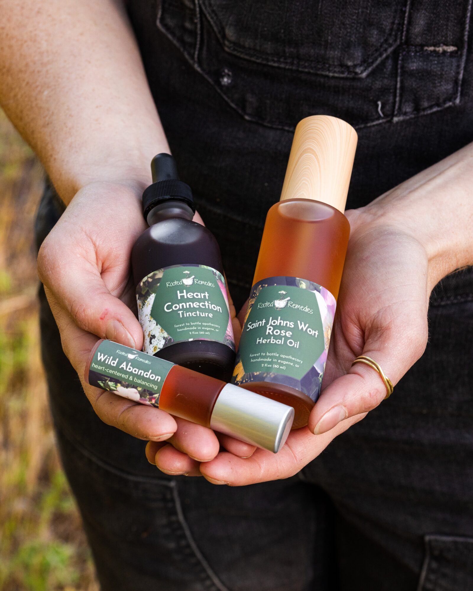 Person holding three bottles and a tube of herbal products outdoors, with a black shirt and jeans visible.