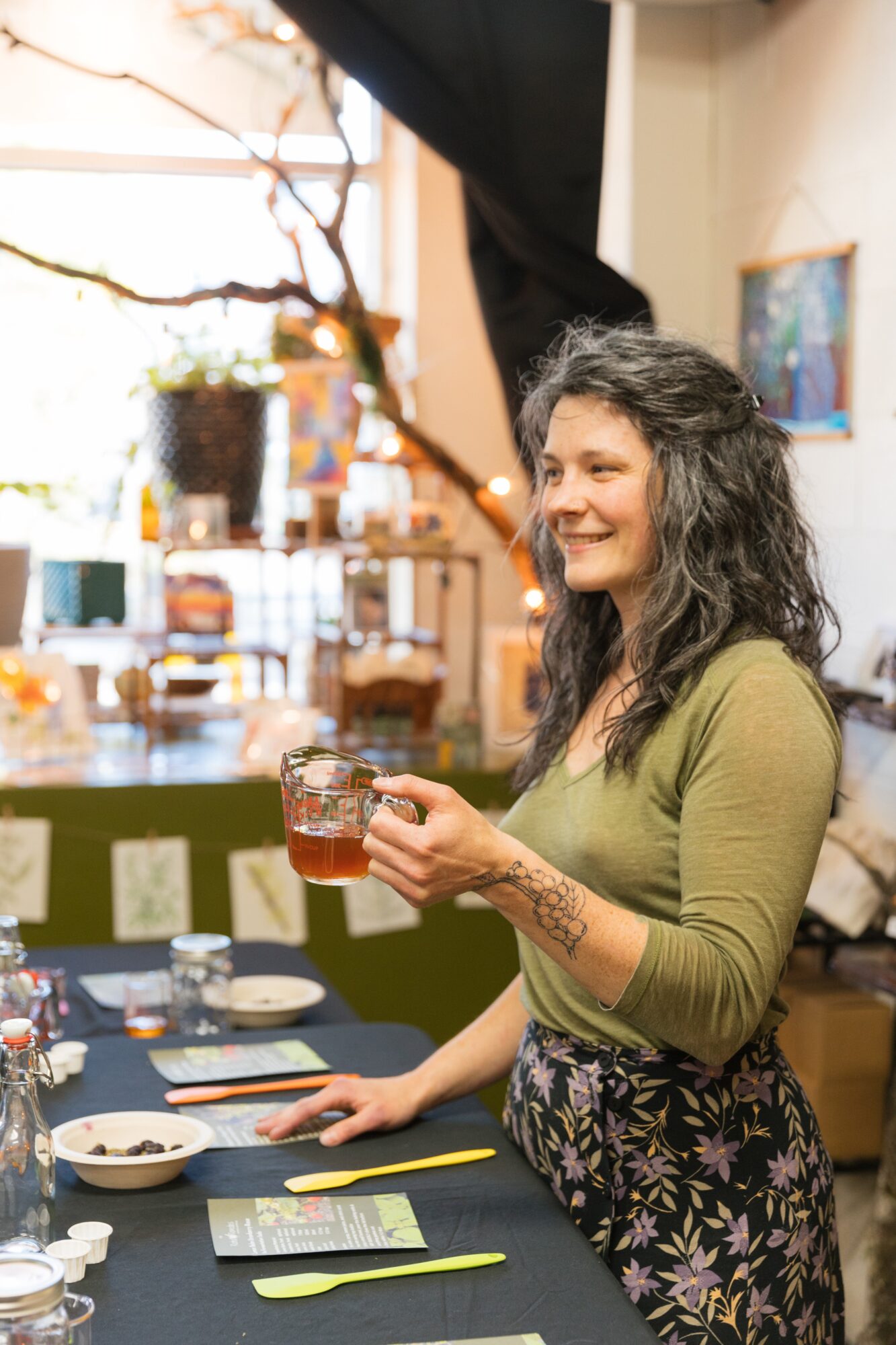 Woman with curly hair smiling, holding a glass, standing at a table in a cozy indoor space.
