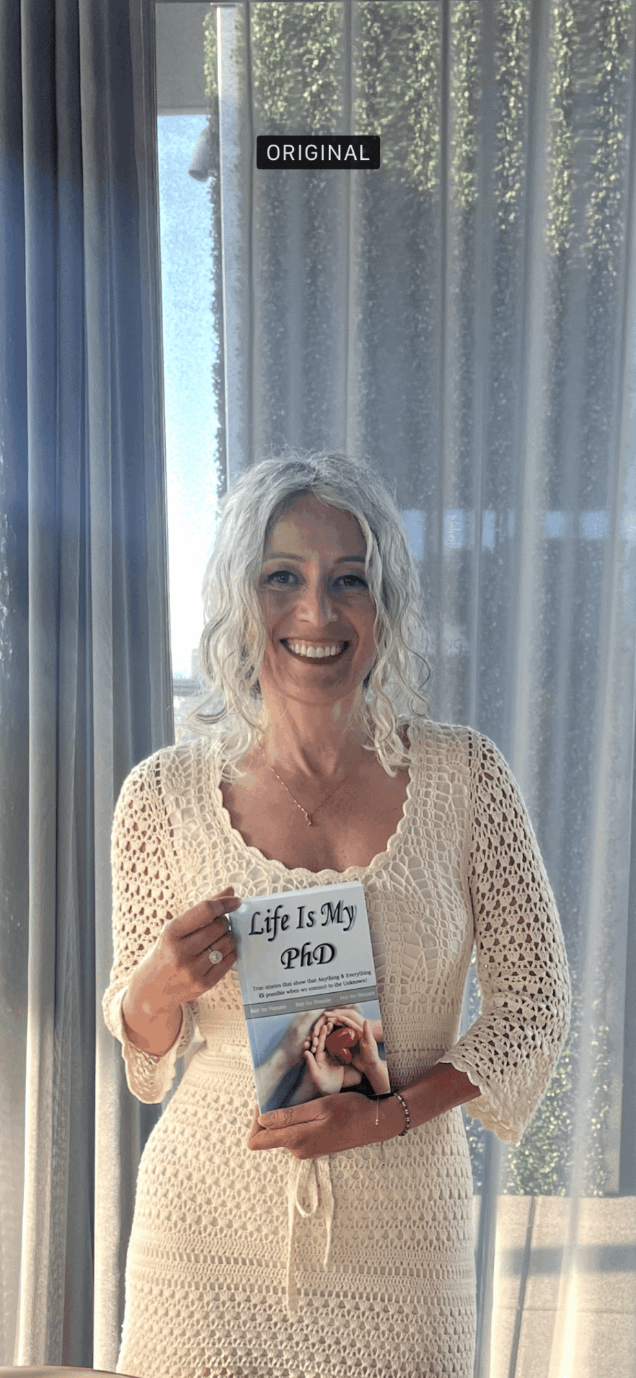 Woman with gray hair smiling, holding a book, standing in front of a window with curtains.