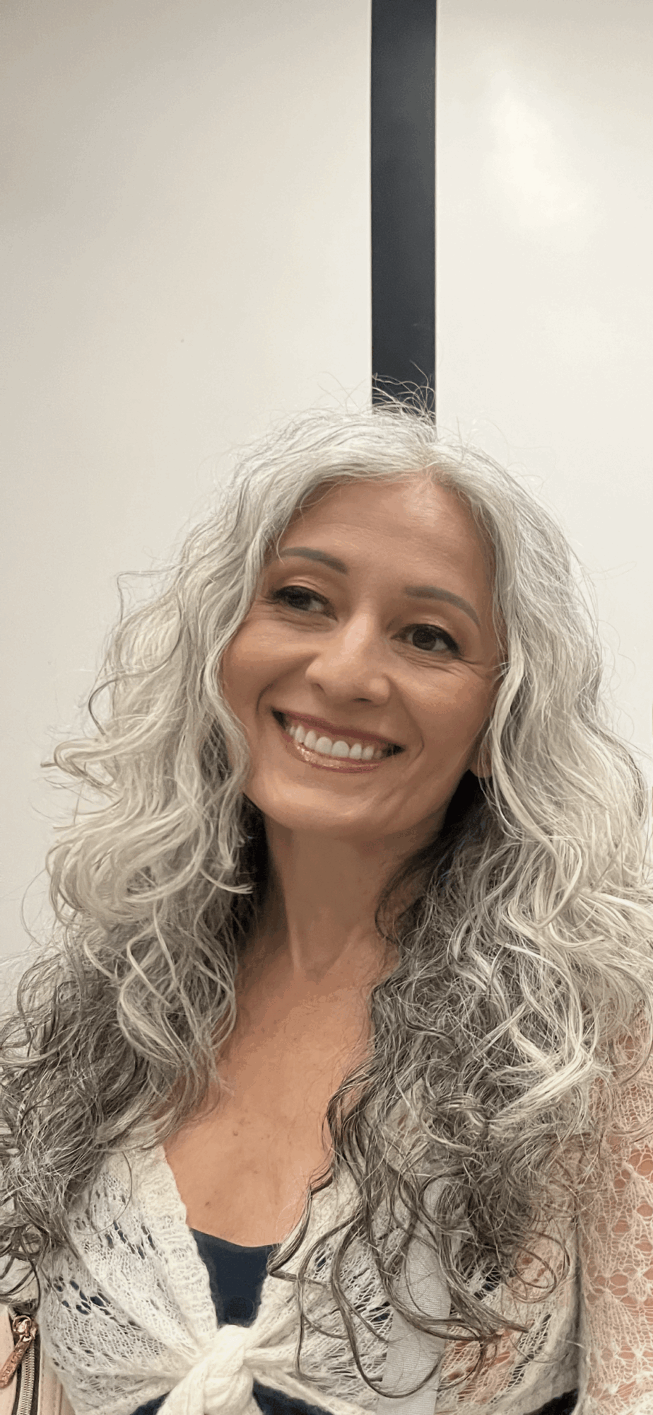 Smiling woman with long, curly gray hair, wearing a light-colored top, standing indoors against a plain background.