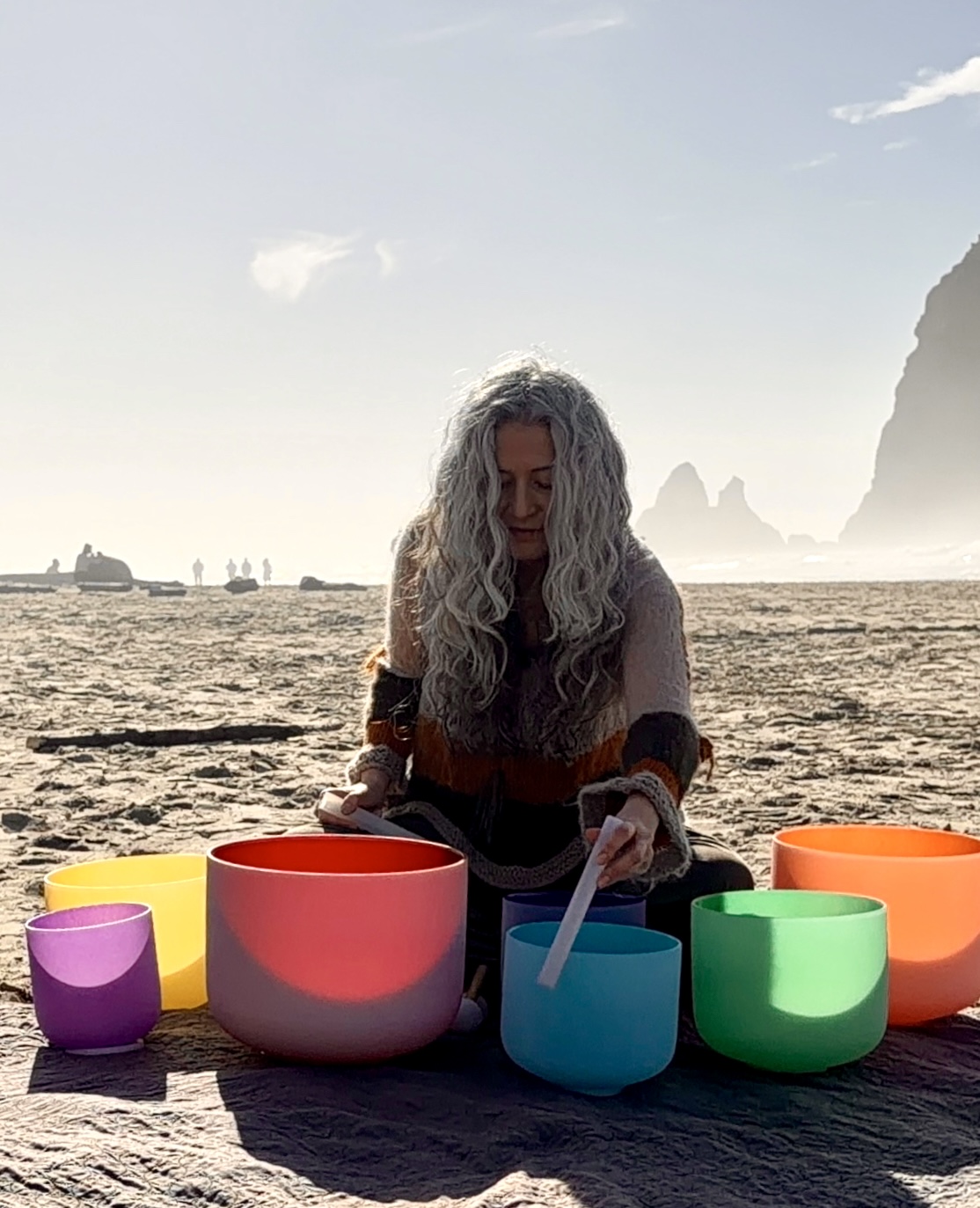 Person with long curly hair playing singing bowls on a beach with rocks and cliffs in background.