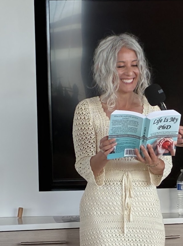 Smiling woman with curly gray hair reads a book aloud into a microphone in a room with a black screen behind her.