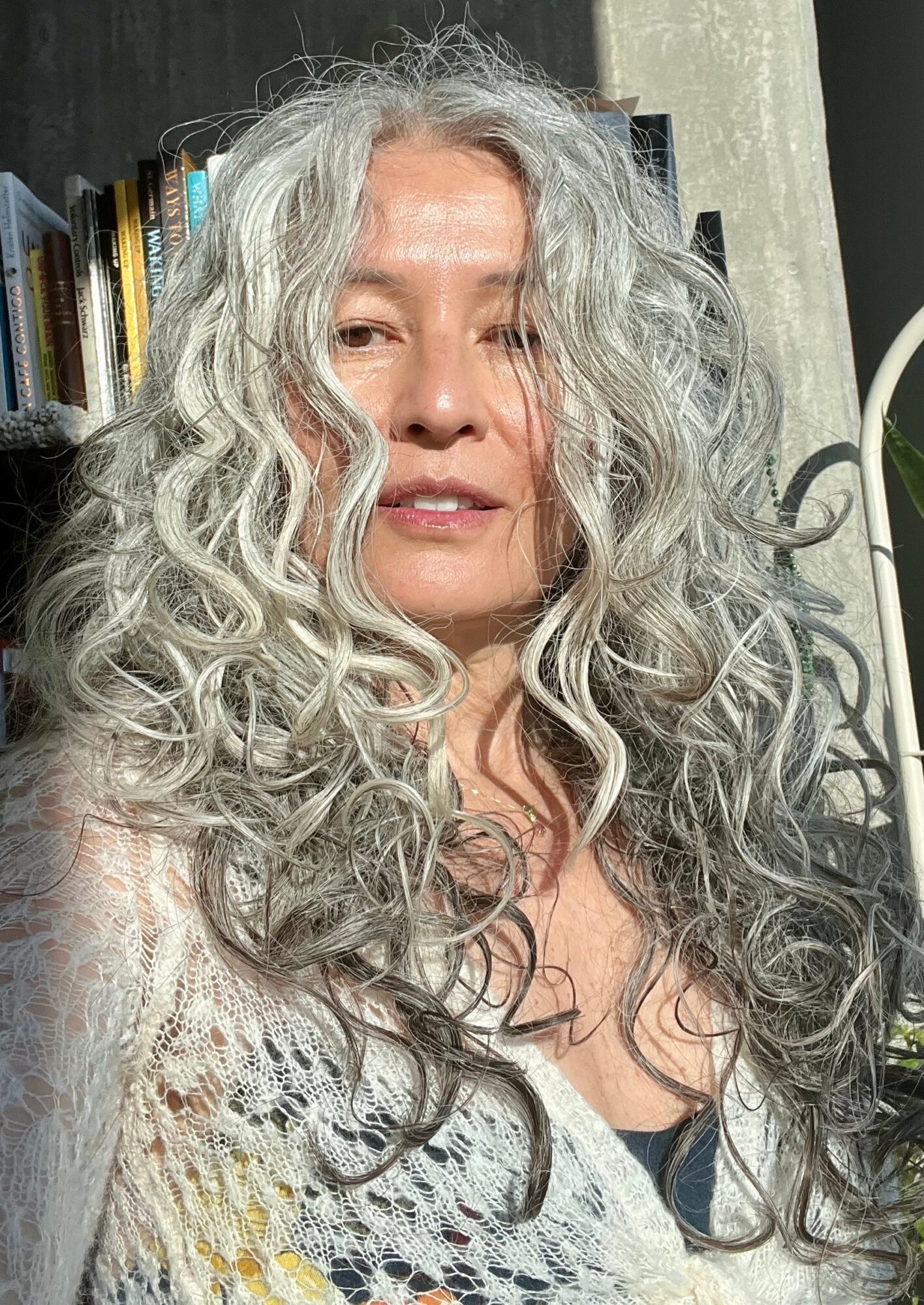 Woman with long, curly, gray hair, wearing a light-colored top, in front of a bookshelf and a curtain.