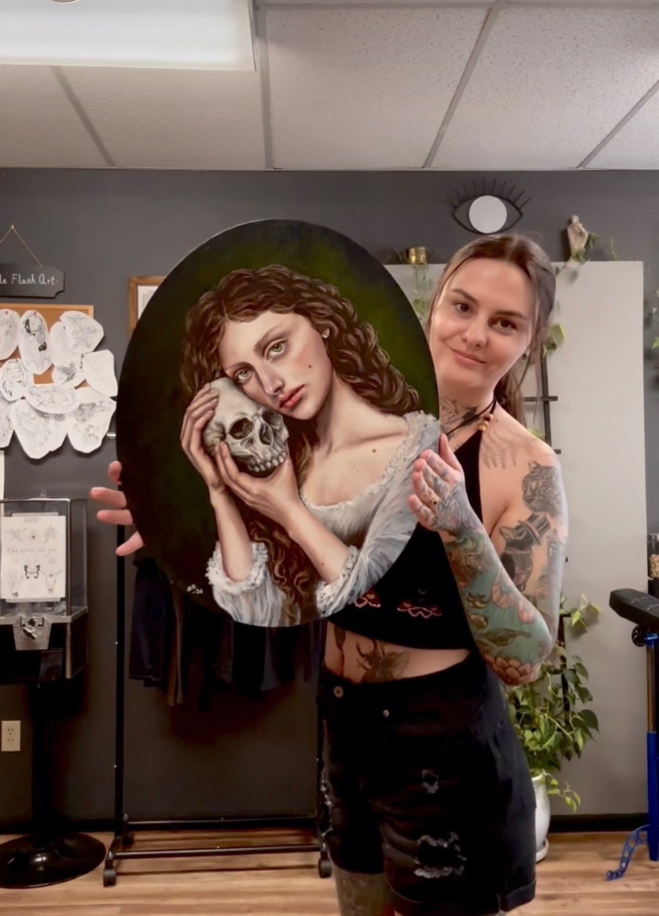 Young woman with tattoos holding a painted oval artwork of a woman with curly hair and skull, smiling in a room.