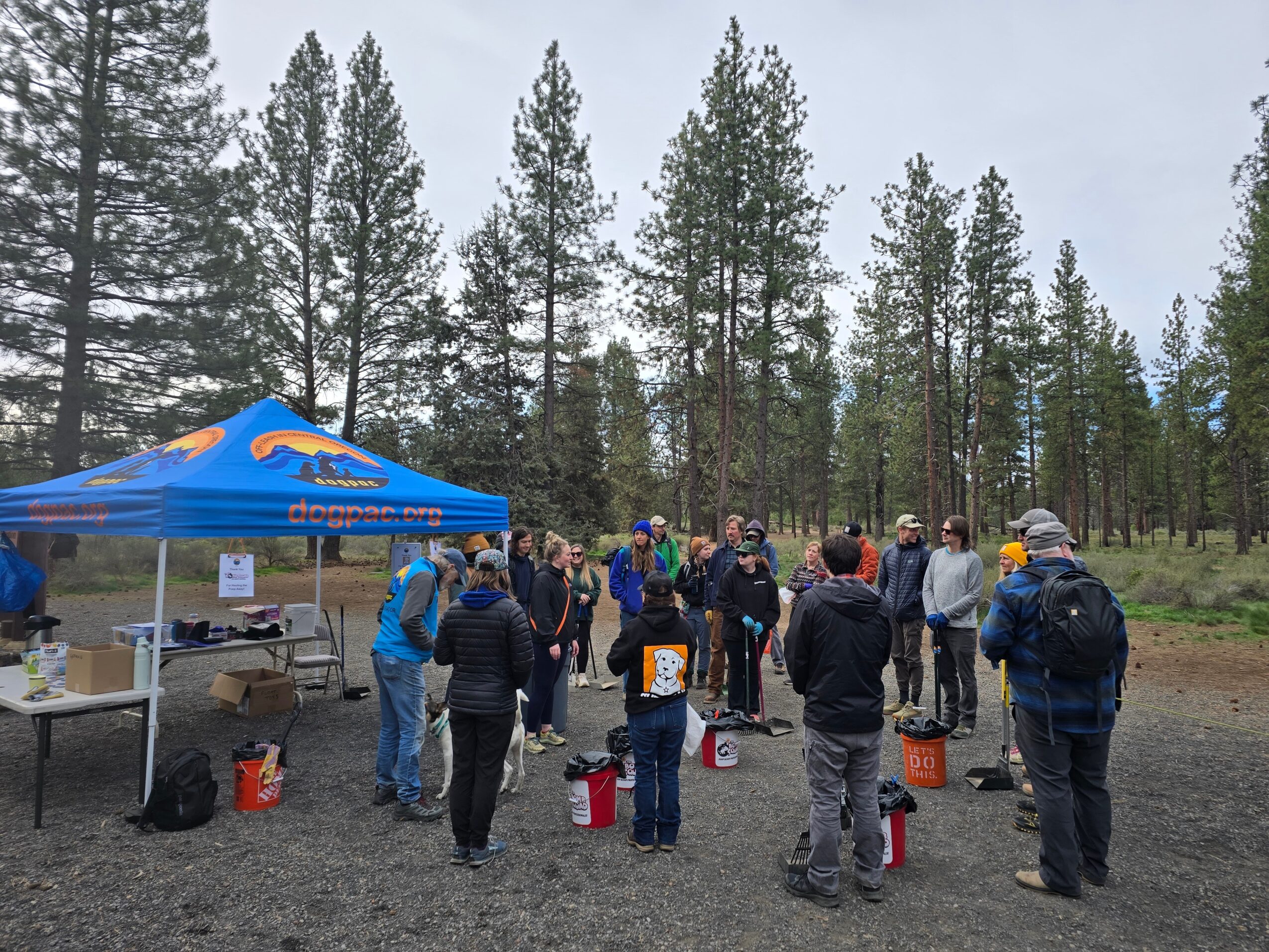 Group of people gathered outdoors near a blue tent in a forested area with tall trees, some holding buckets or containers.
