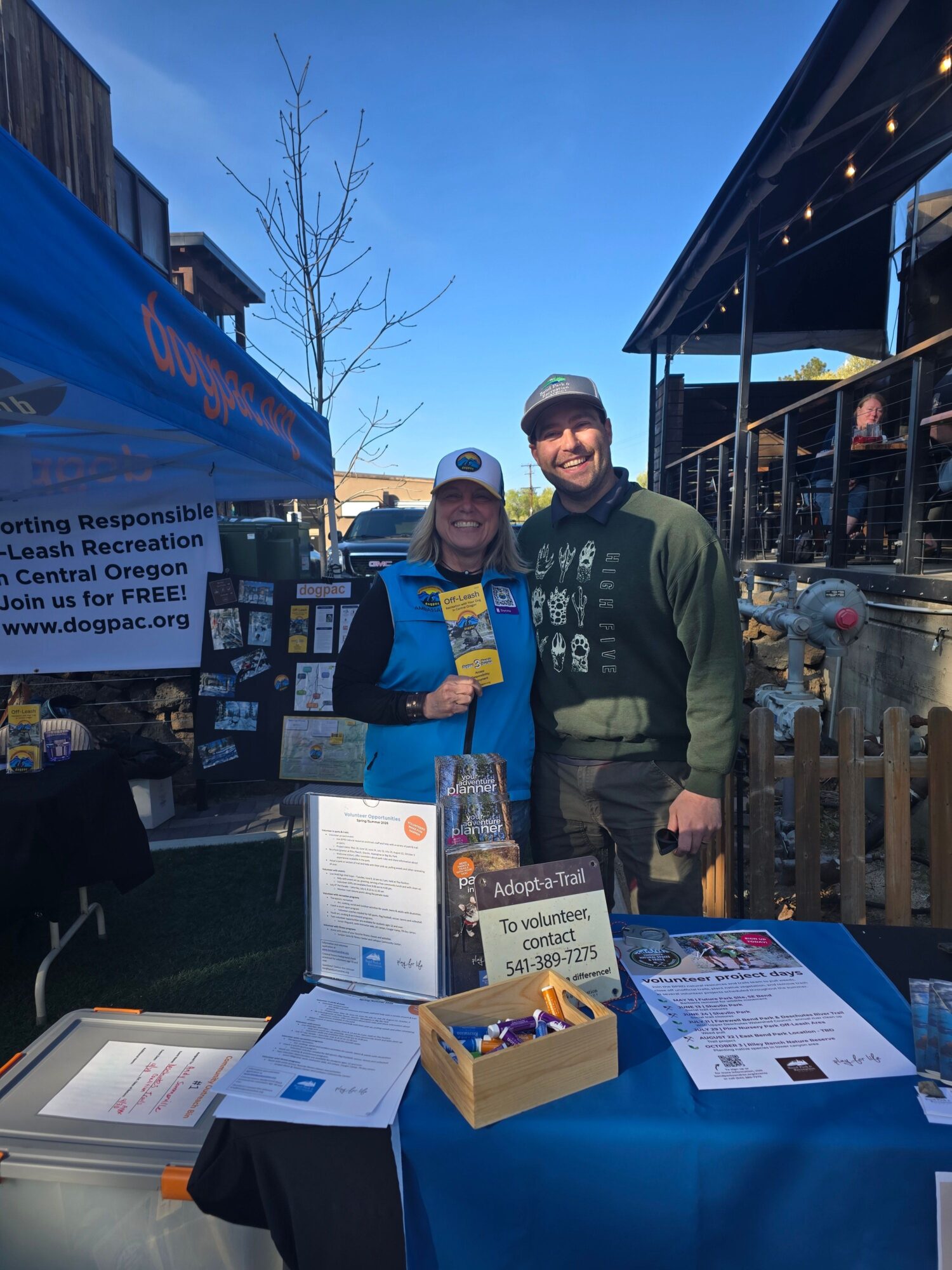 Two people standing behind a table with informational materials at an outdoor event during evening. One person wears a blue jacket, the other a green hoodie. A third person is partially visible on the right. The table has flyers, a box, and a sign. Background shows a building and a tree.