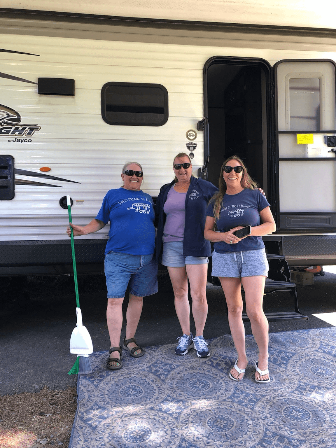 Three people stand in front of a white RV, smiling, with a broom leaning against the RV, on a patterned outdoor rug.