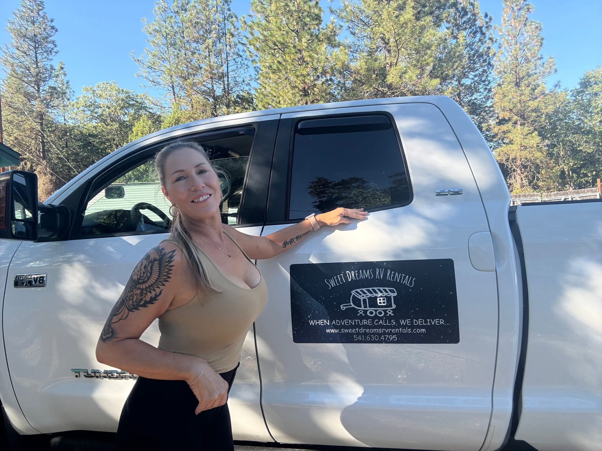 Woman with tattoos smiling and pointing at a sign on a pickup truck's door, outdoors with trees and blue sky.