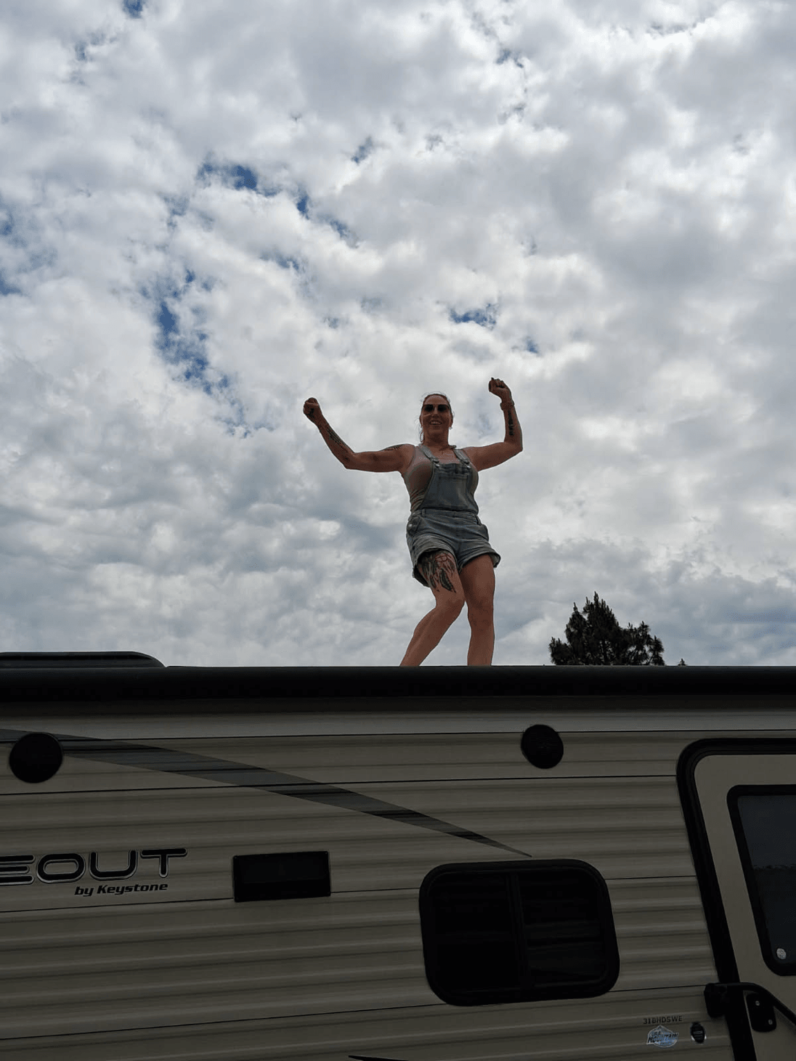 Person standing on a roof with arms raised, sky with clouds in background, tree visible below, smiling, wearing sunglasses and shorts.