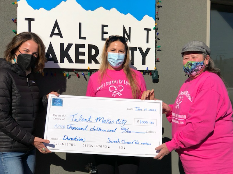 Three women holding a large check outdoors, with a sign reading 'Talent Maker City' behind them. All wear masks.