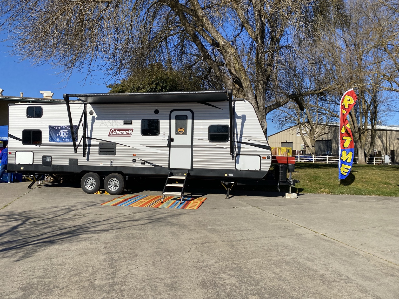 Travel trailer parked on a lot with a colorful mat, tree in background, and a flag with the word 'LOVE'.