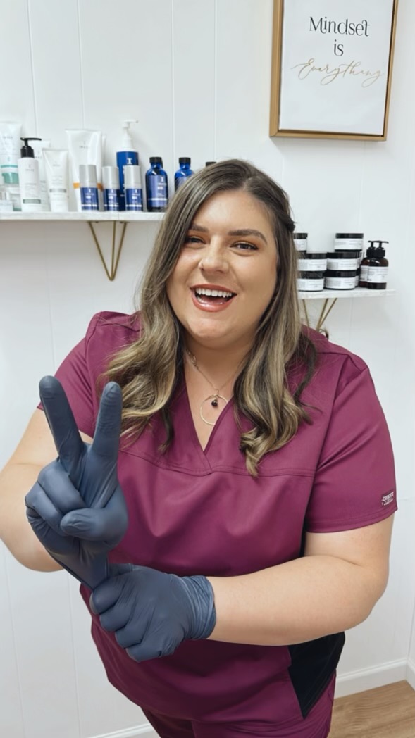 Smiling woman in purple scrubs making a peace sign with gloved hand, shelves with skincare products behind her.