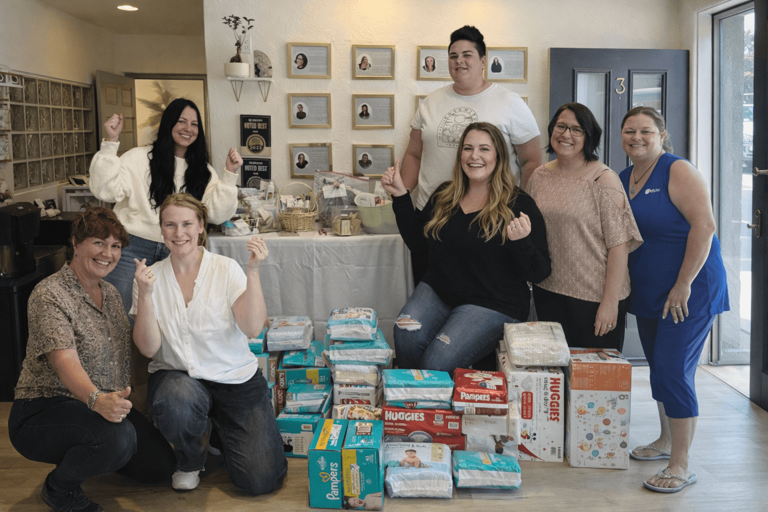 Group of seven women and one man standing and sitting behind a table with diapers and gifts, smiling indoors.