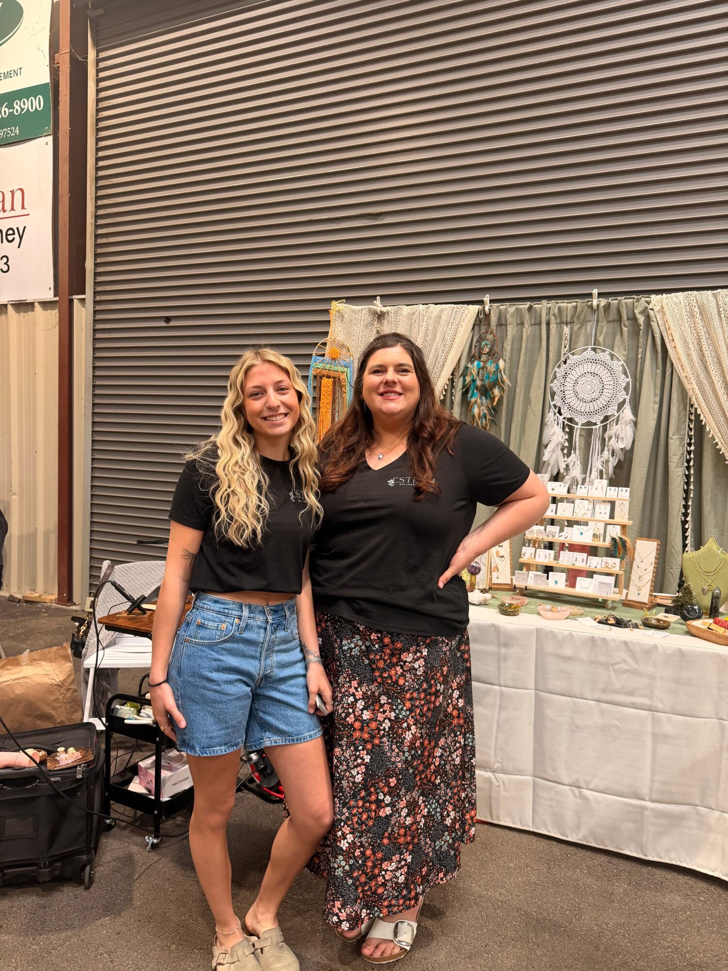 Two women standing indoors, one in a black top and floral skirt, the other in a black top and denim shorts, smiling.