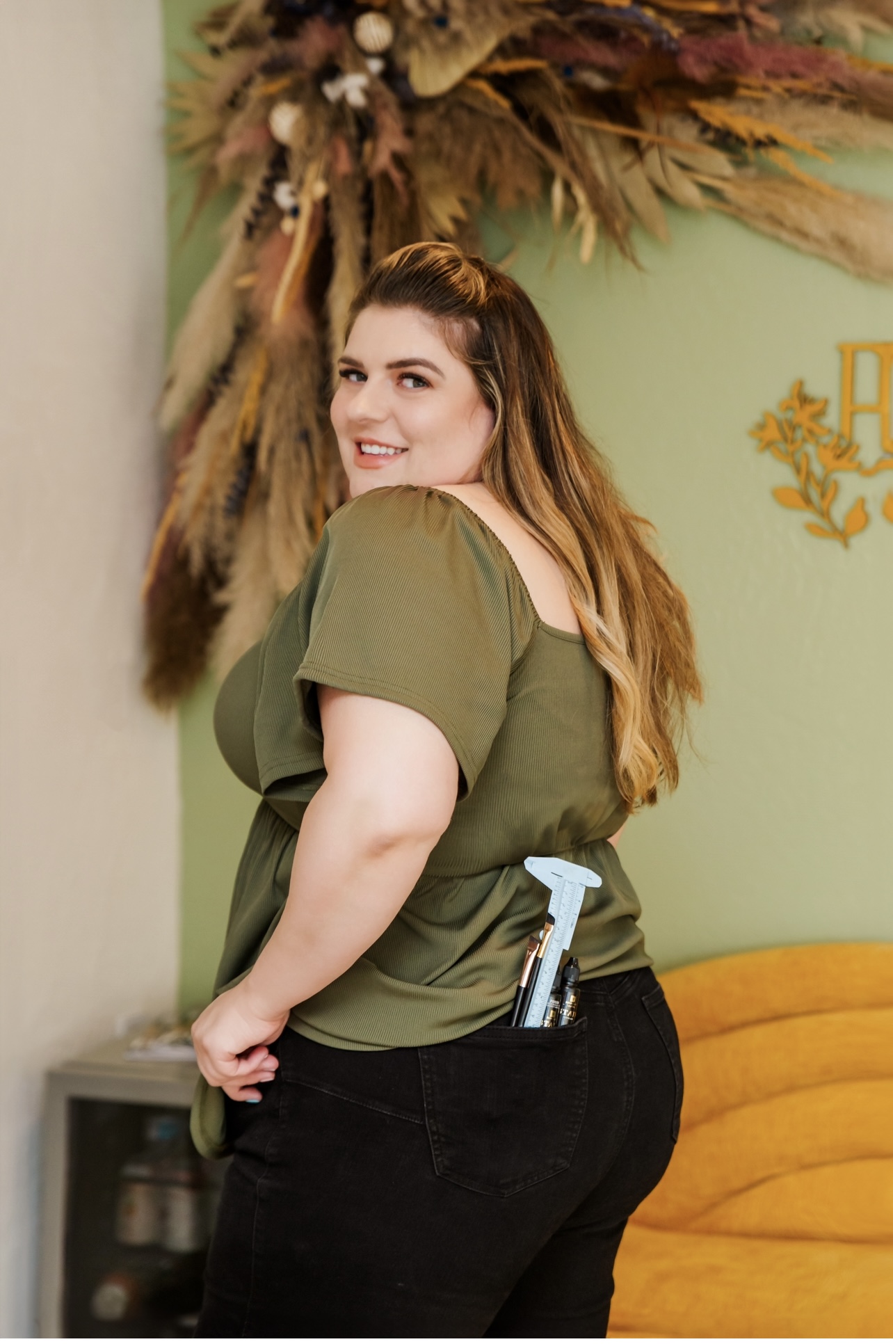 Smiling woman with long hair in an olive green top and black pants, standing indoors near a yellow couch and decorative wall art.
