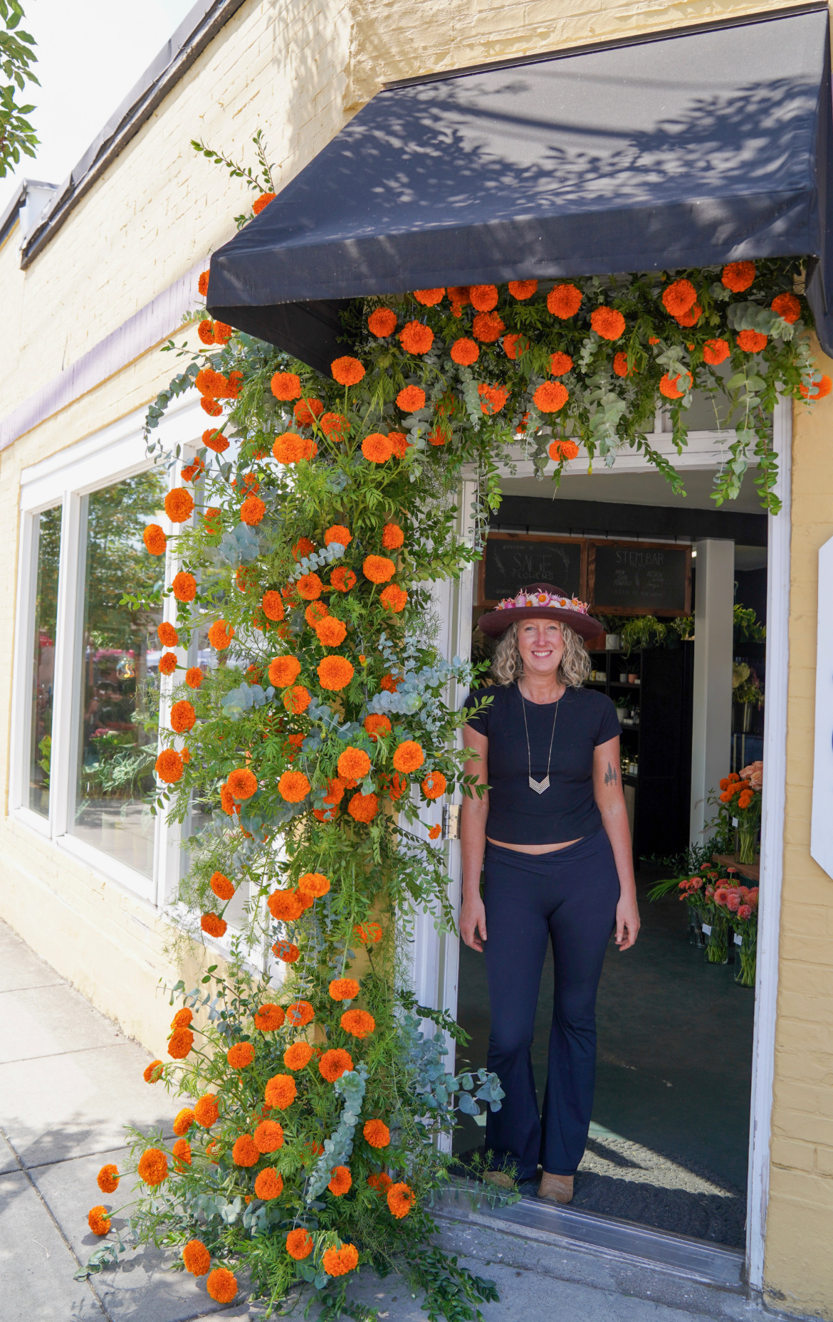 Woman standing in doorway decorated with orange and white flowers, wearing a hat and dark clothing.