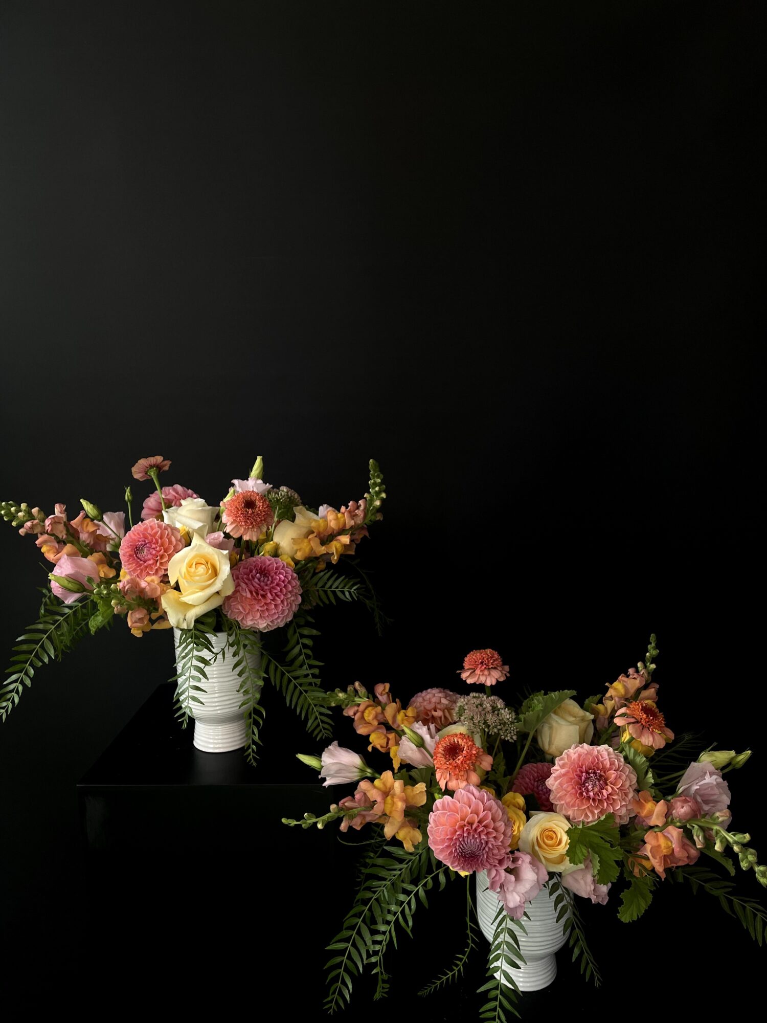 Two floral arrangements in white vases with pink, yellow, and orange flowers and green foliage against a black background.