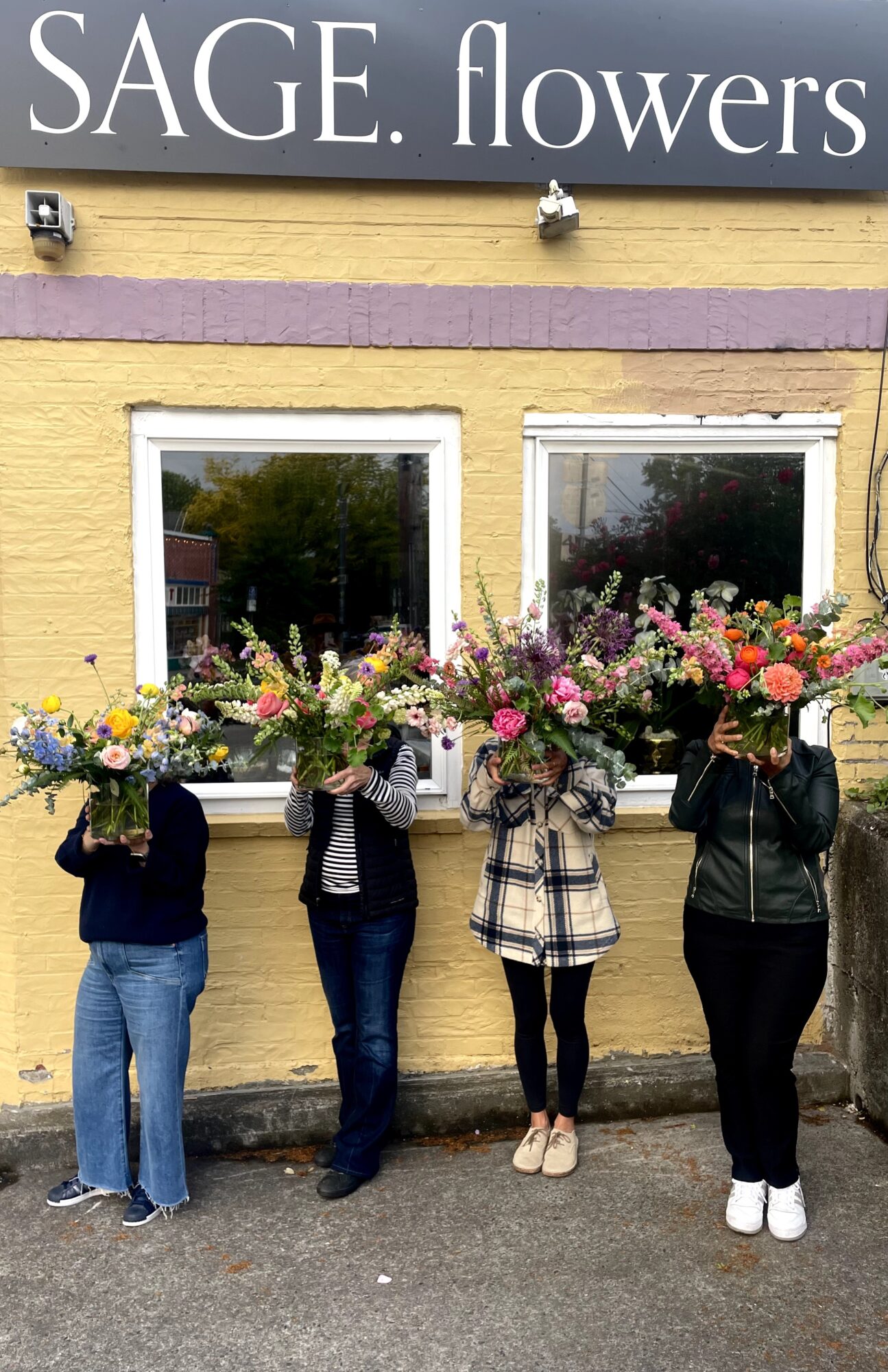 Four people holding flower bouquets in front of a yellow building with two windows, with a sign reading 'SAGE flowers' above.