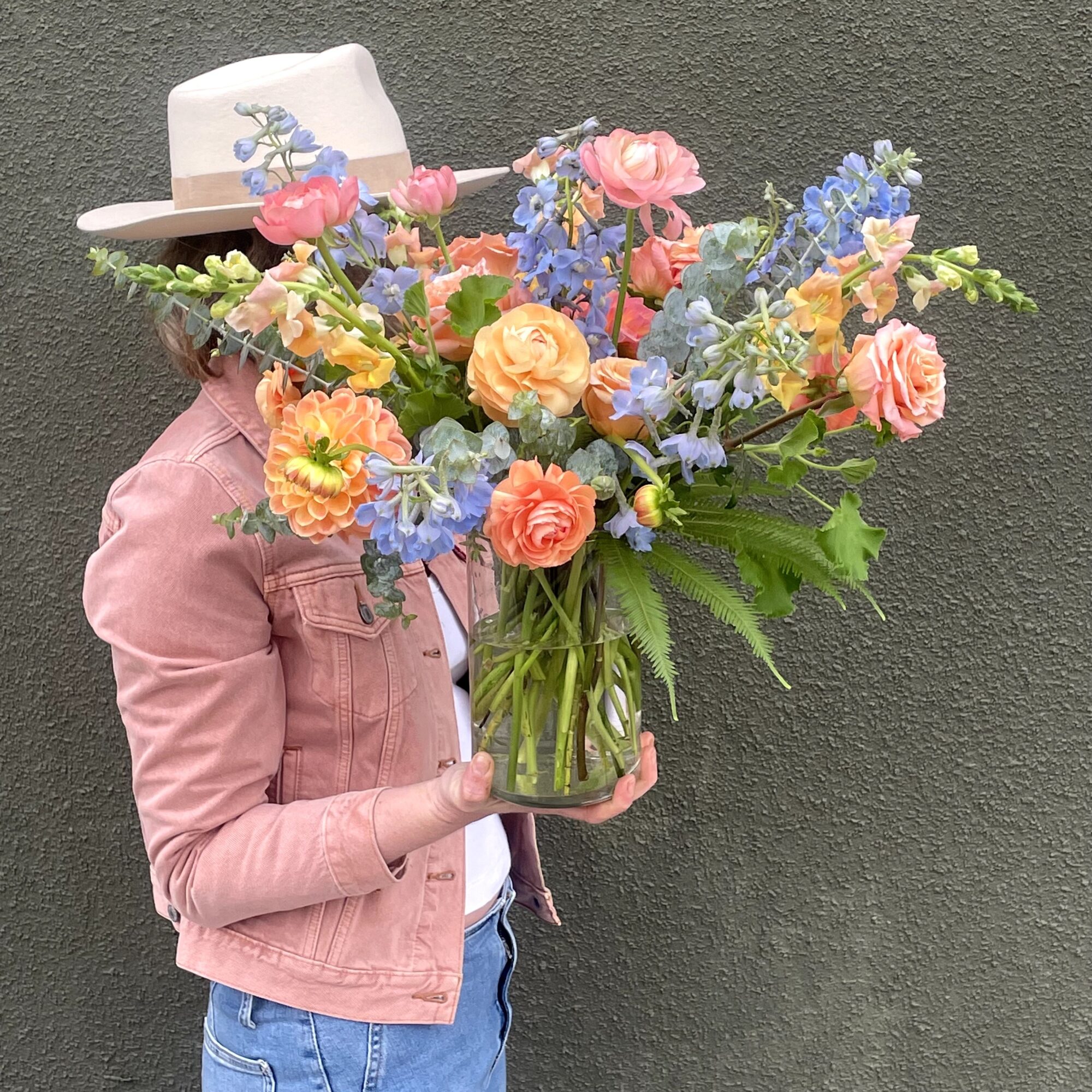 Person holding a large bouquet of colorful flowers in front of a dark wall, wearing a hat and pink jacket.
