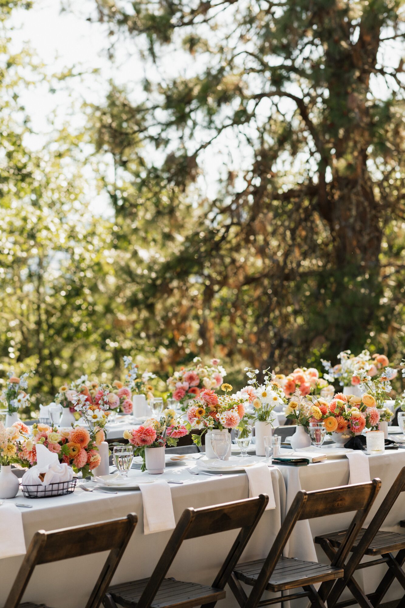 Outdoor table set with floral arrangements and chairs, surrounded by trees and sunlight filtering through leaves.