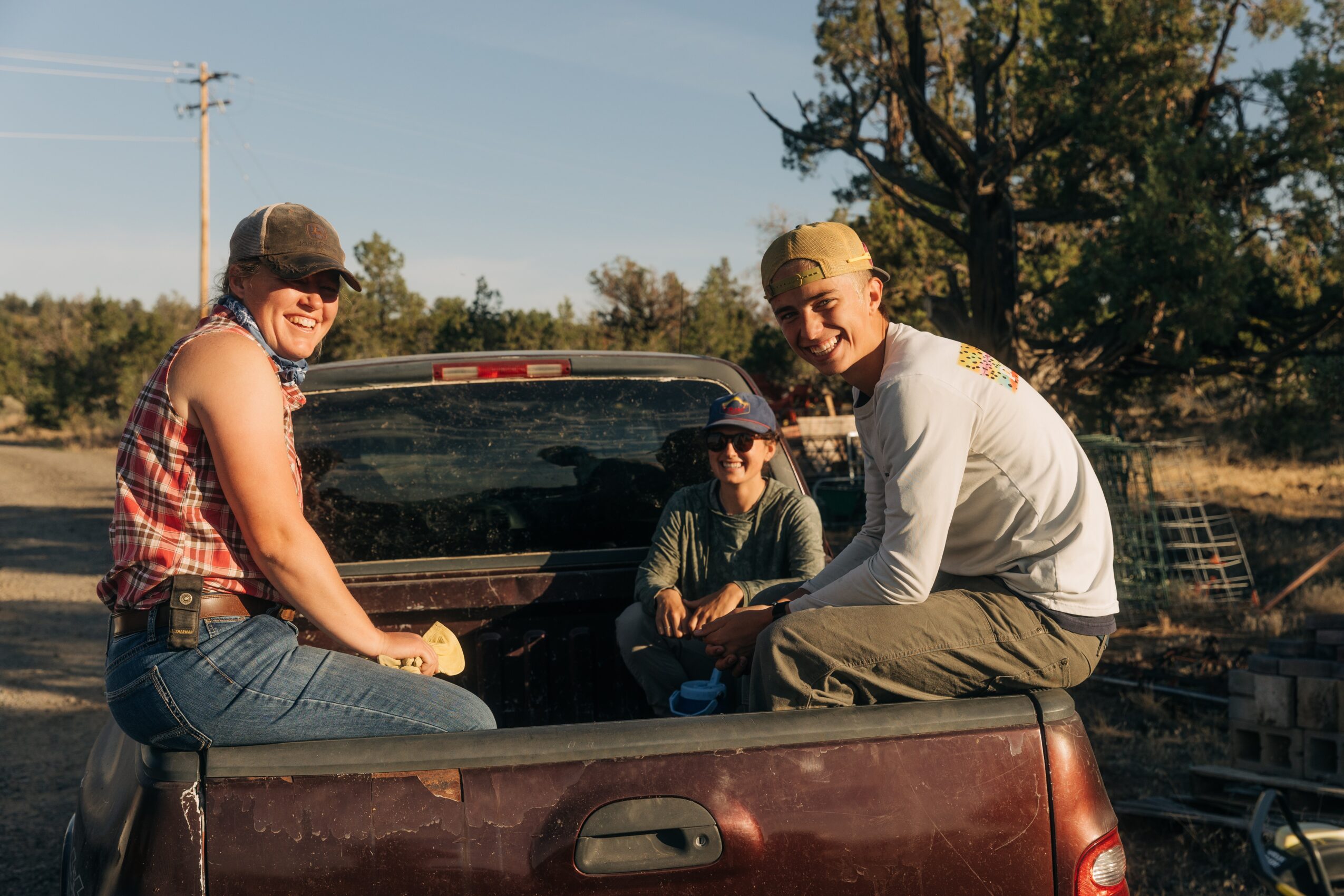 Three people sitting on the back of a pickup truck outdoors, smiling, with trees and a clear sky in the background.