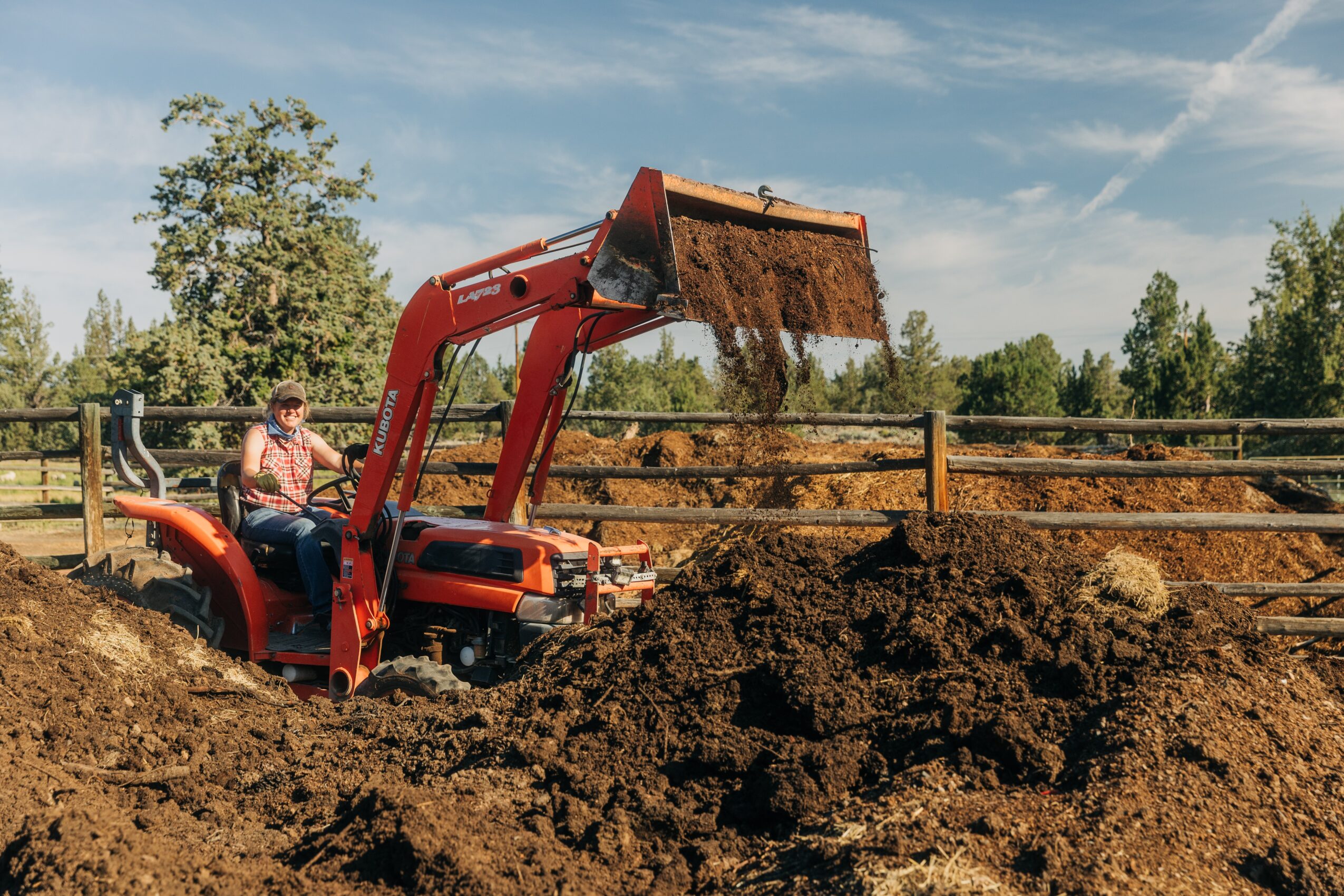 Person operating a small orange tractor with a front loader, moving dirt in an outdoor fenced area, trees in background.