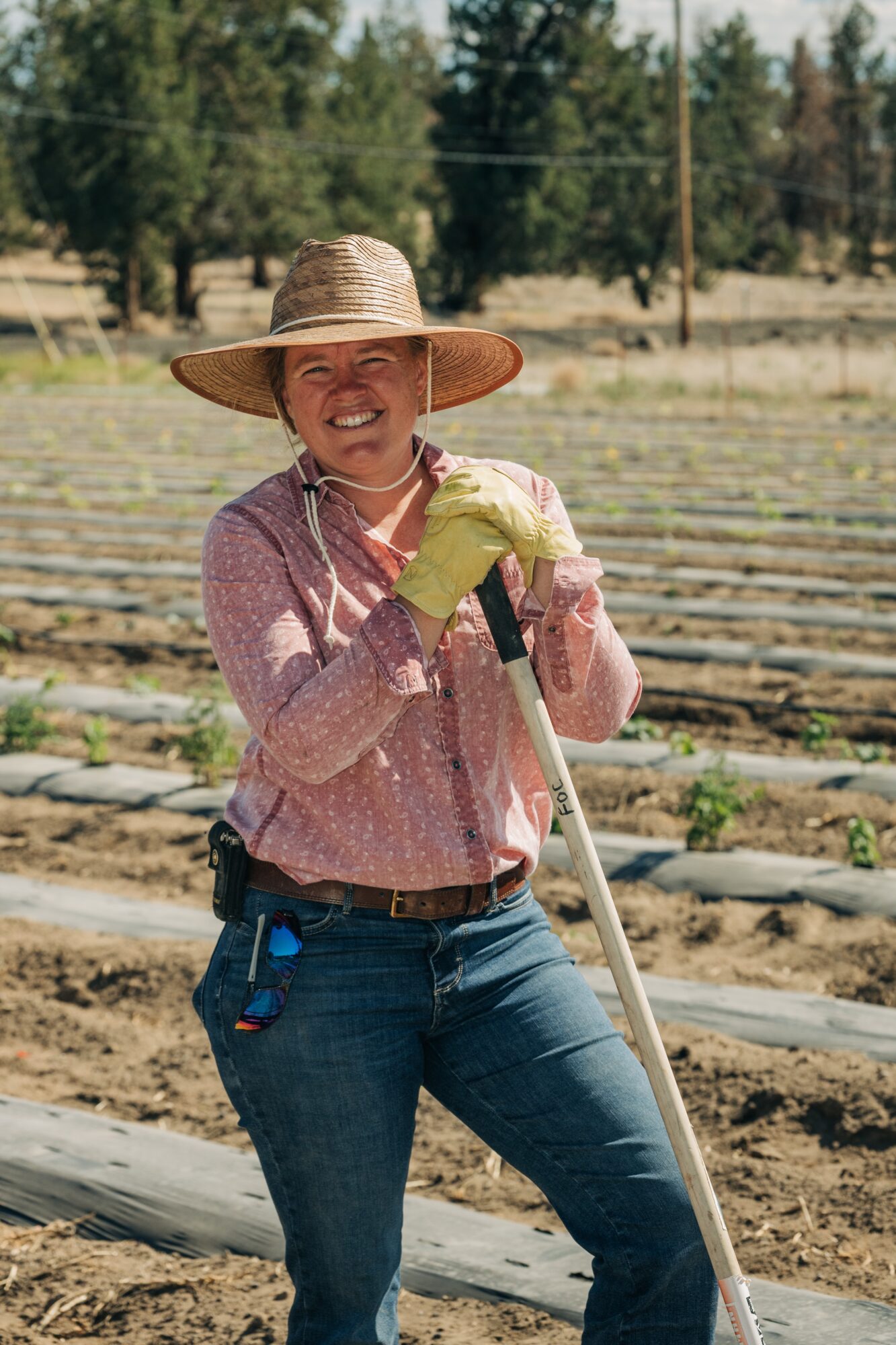 Woman wearing a wide-brimmed hat and gloves, smiling outdoors in a garden or farm setting.