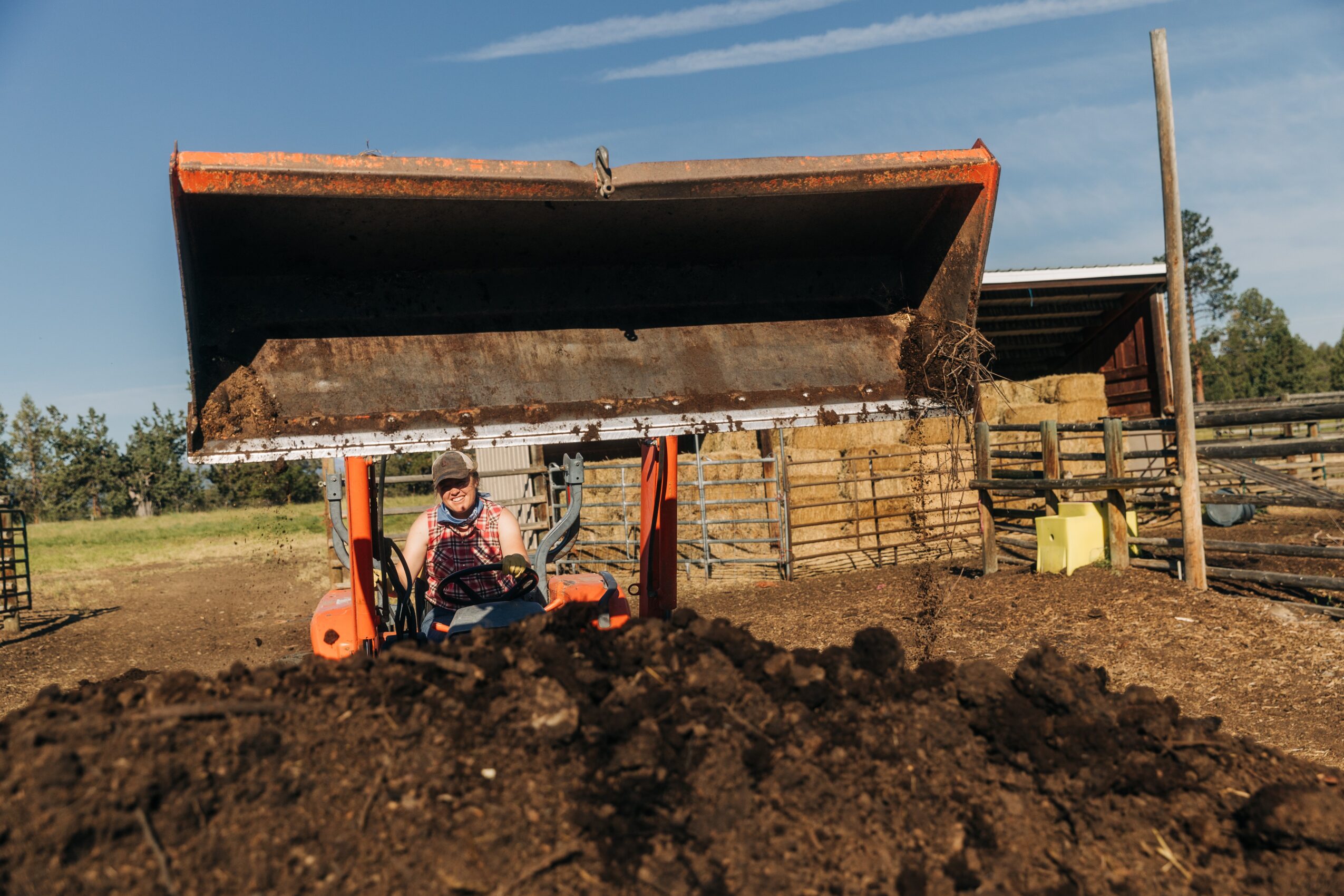 Person operating a tractor with a large raised scoop of dirt, outdoors on a farm or rural area, with a fence and trees in background.