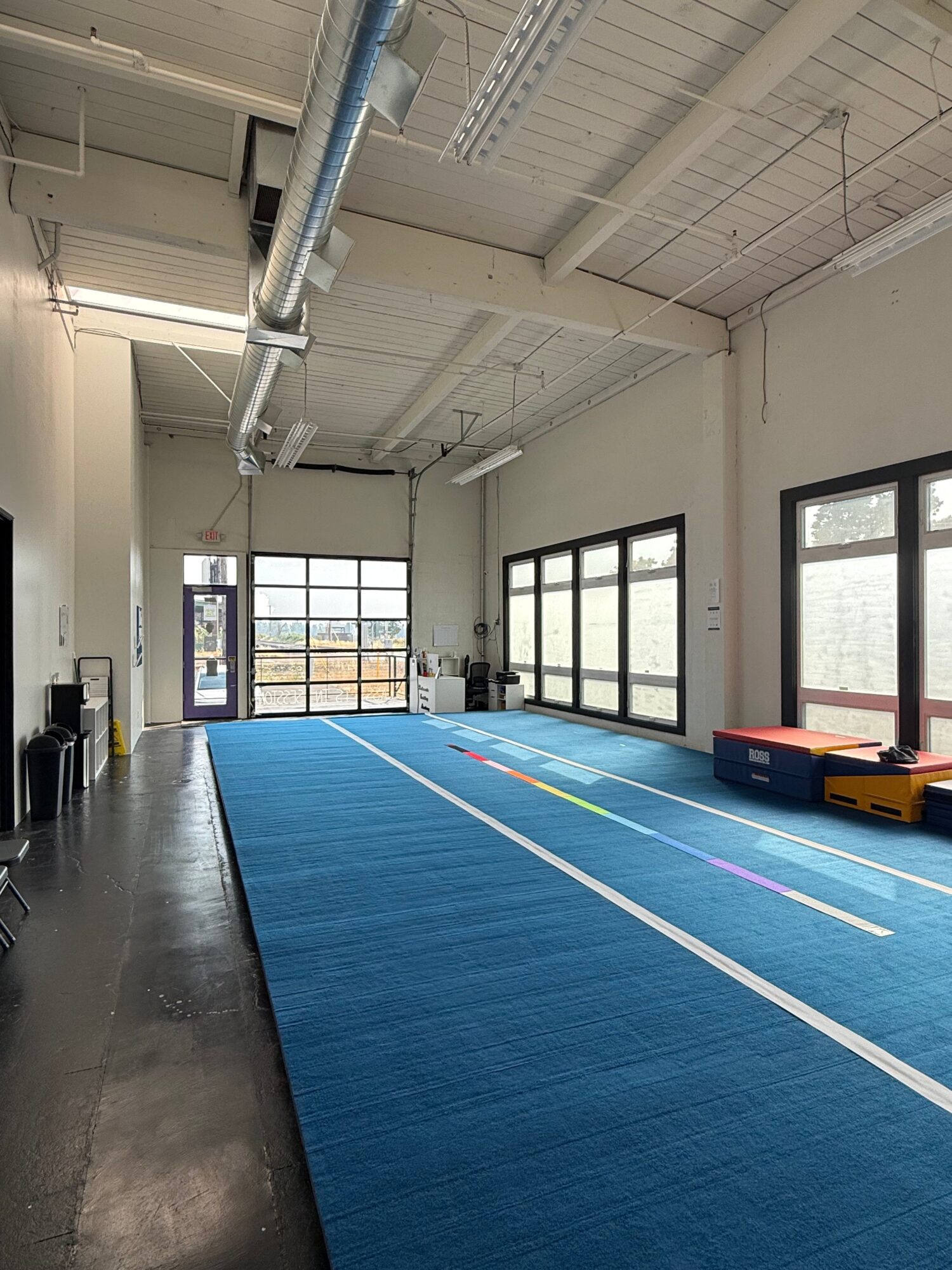 Indoor gym with blue mat, windows, and equipment, viewed from the side, with natural light coming in.