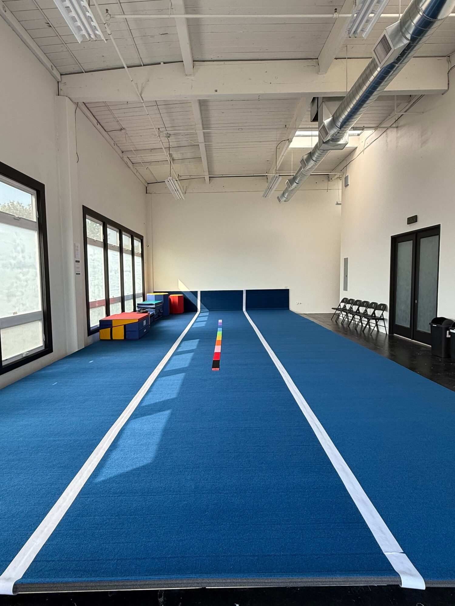Indoor gym with blue mat, white lines, and windows, viewed from the side. Equipment and a door are visible in the background.