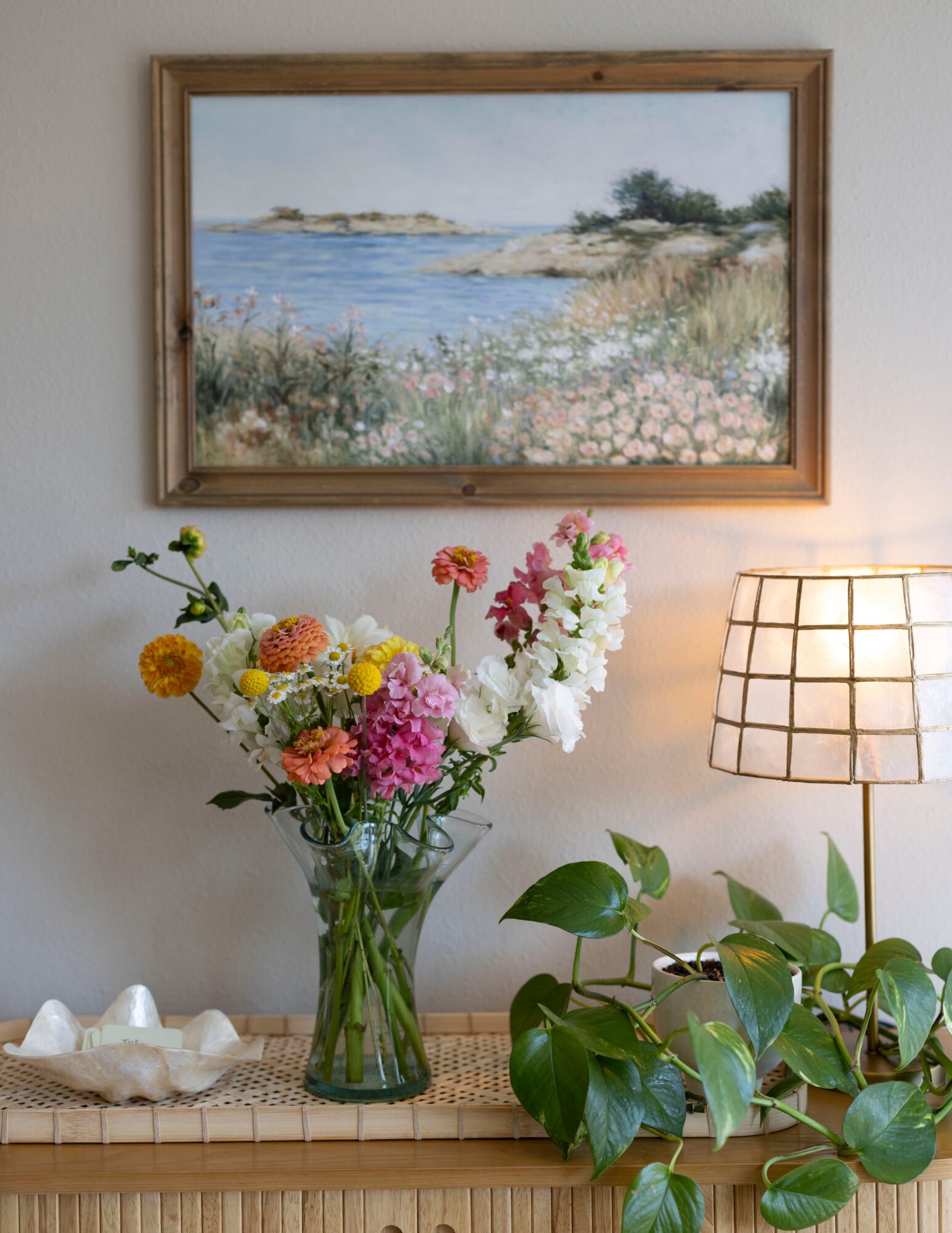 A room with a landscape painting, a vase of colorful flowers, a table lamp, and a potted plant.