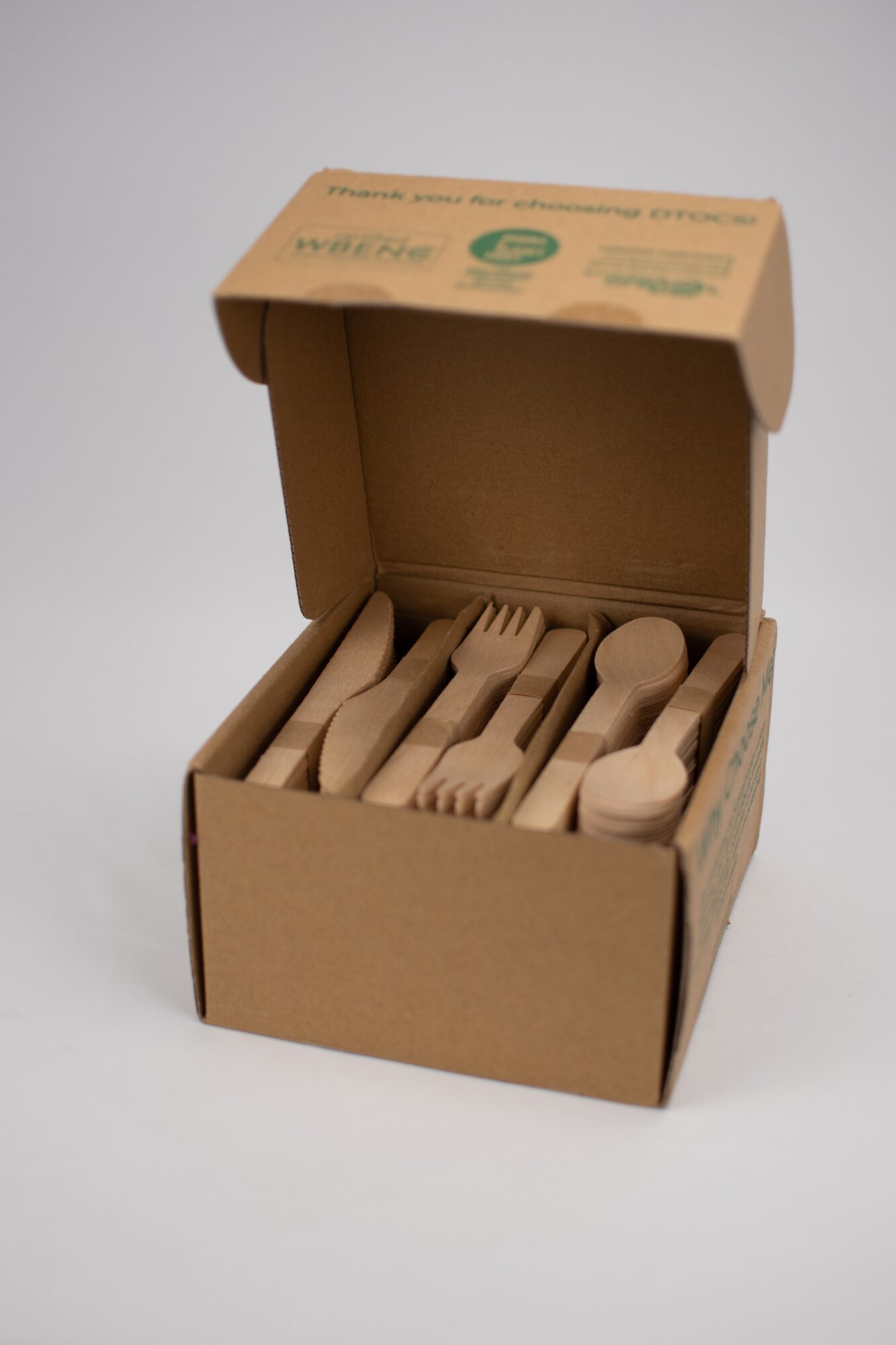 Open cardboard box containing wooden utensils including forks, knives, and spoons, arranged neatly inside. Plain background.