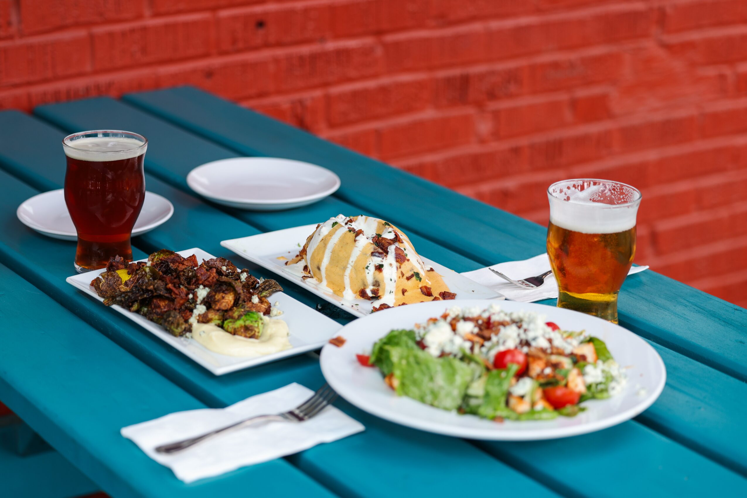 Two plates of food, a salad and a dessert, with two glasses of beer on a blue table against a red brick wall