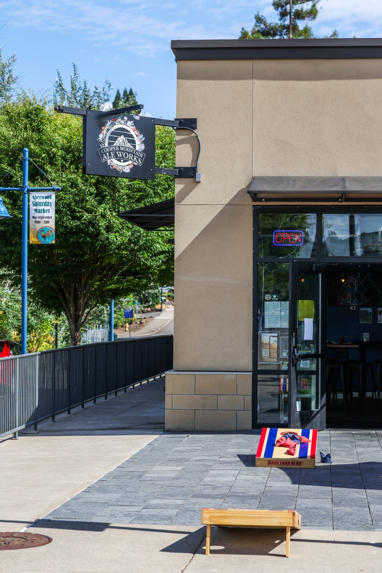 Side of a building with a sign, sidewalk, and trees in the background, sunny day, no people visible.