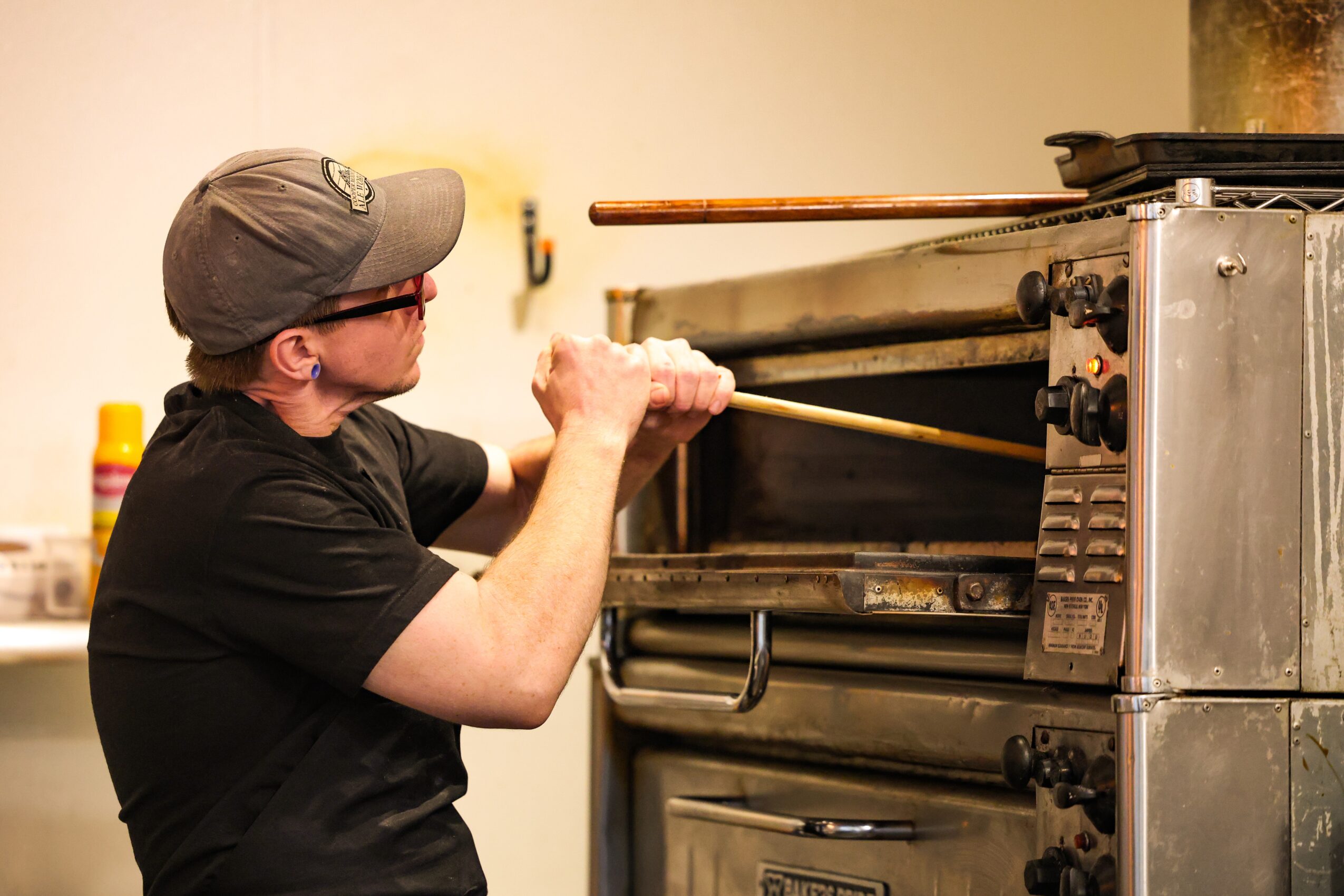Person wearing glasses and a cap using a long tool to open an oven door in a kitchen setting.