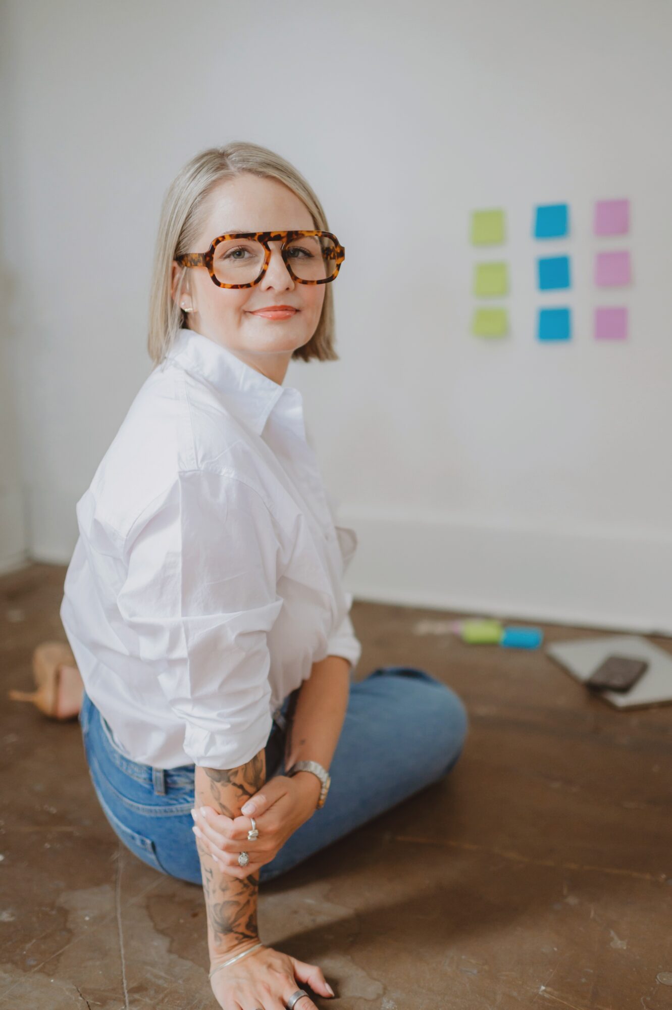 Woman with glasses and tattoos sitting on the floor, white wall with colorful sticky notes behind her.